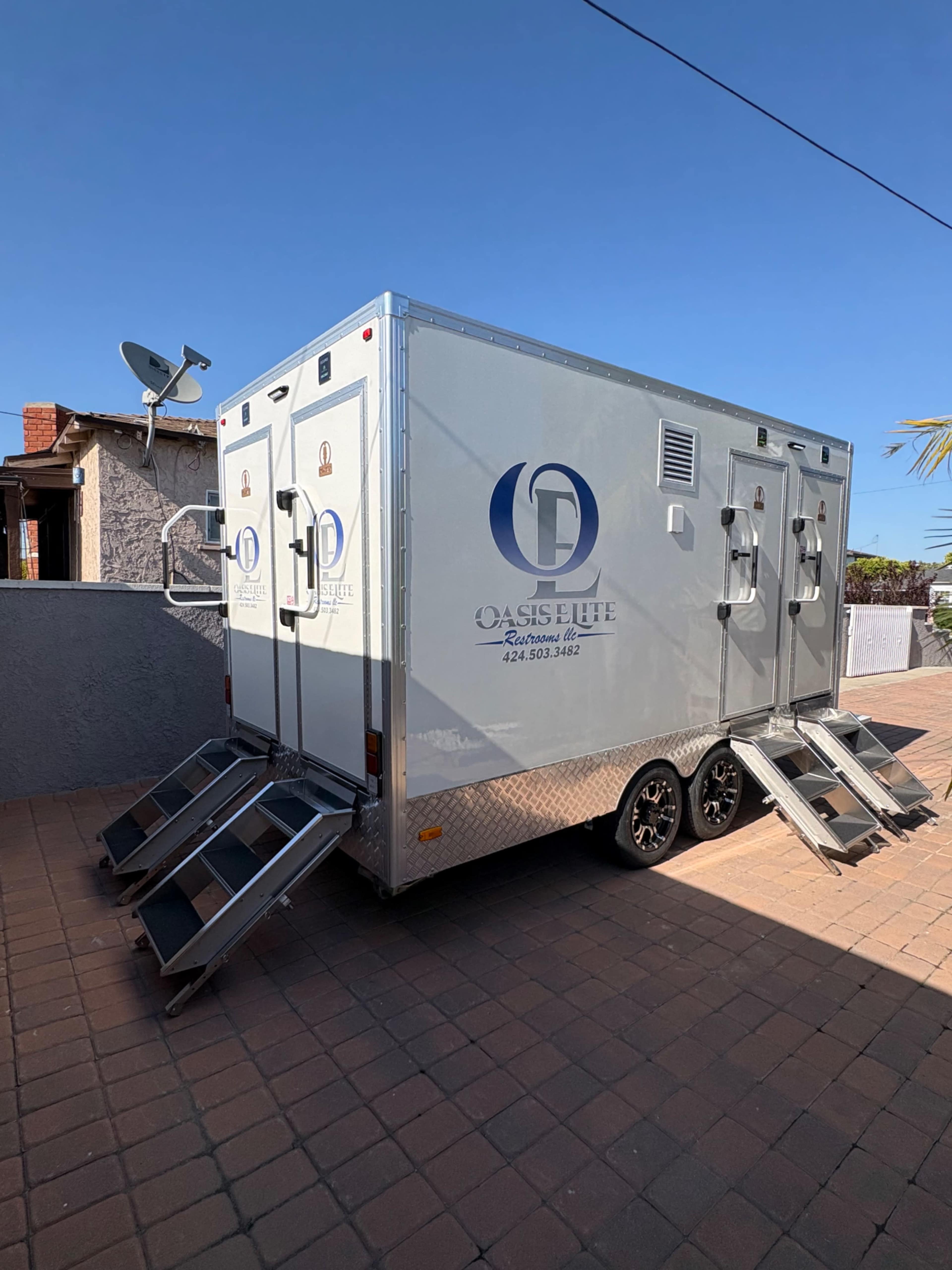 A silver portable restroom trailer with multiple entrances is parked on a brick pathway under a clear blue sky.