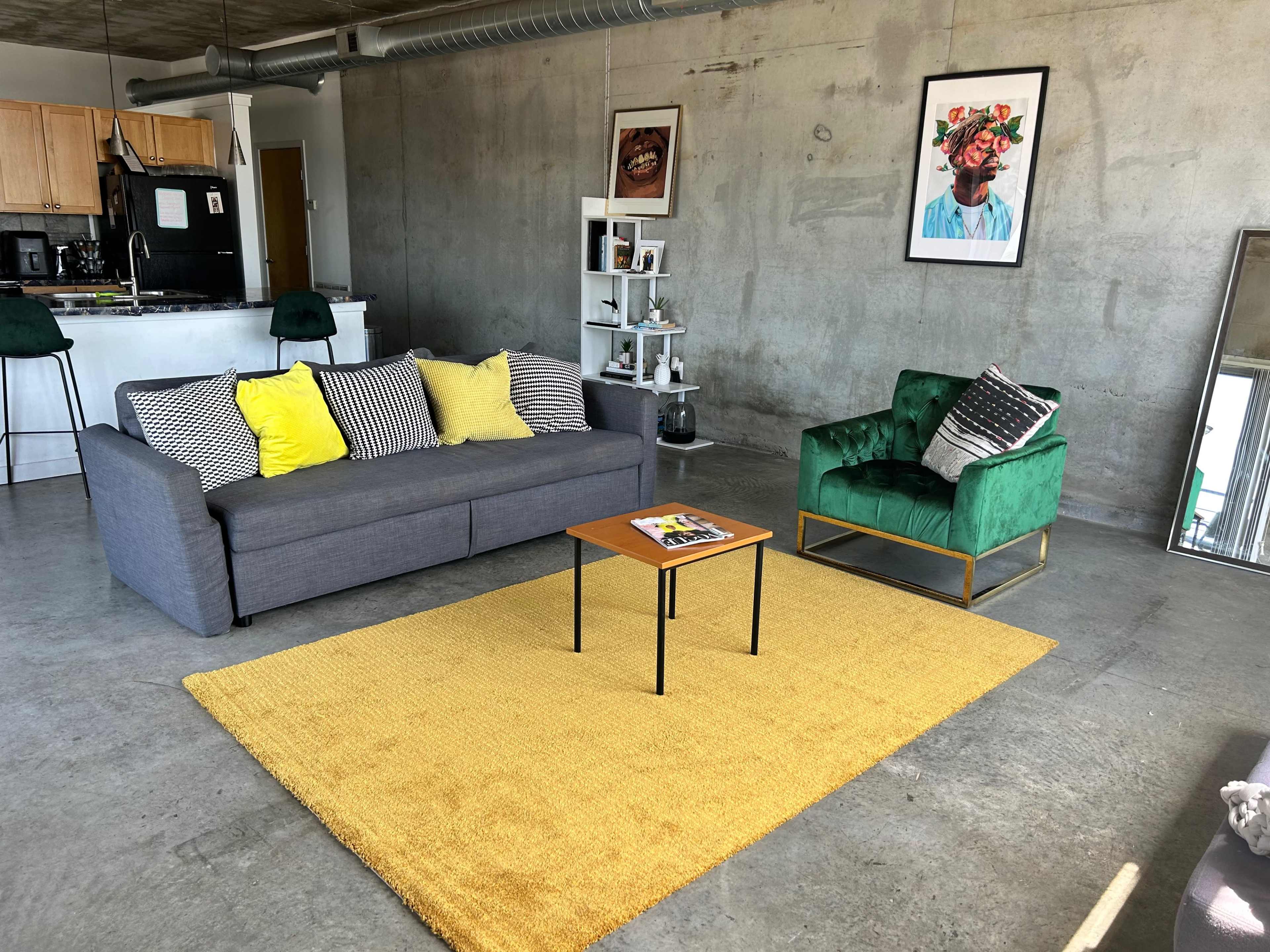 A living room with a gray sofa, green armchair, yellow rug, and a wooden coffee table in front of a concrete wall.