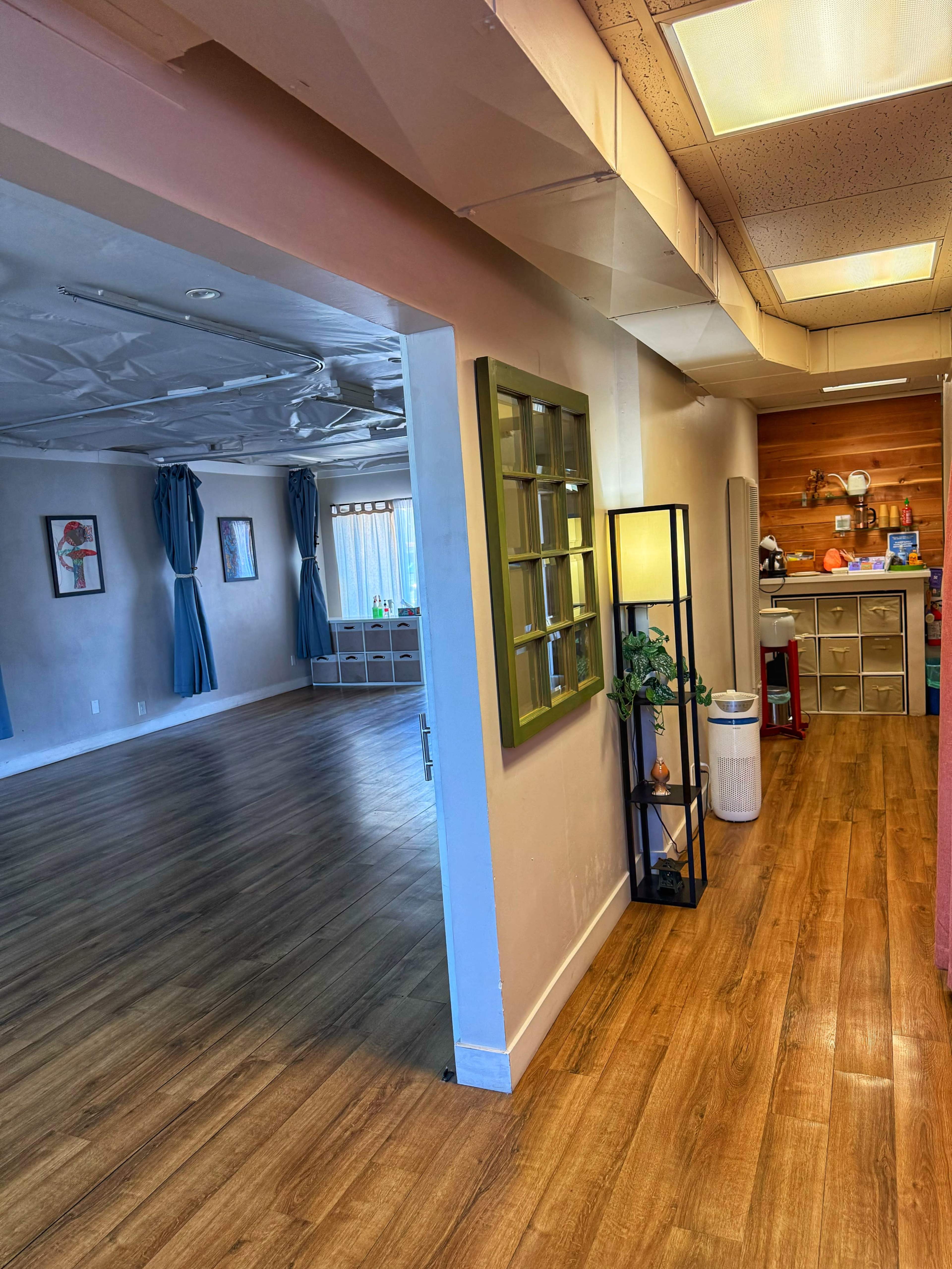 The image shows a hallway leading to two separate rooms, with a green-framed window on the wall and a kitchen area visible in the background.