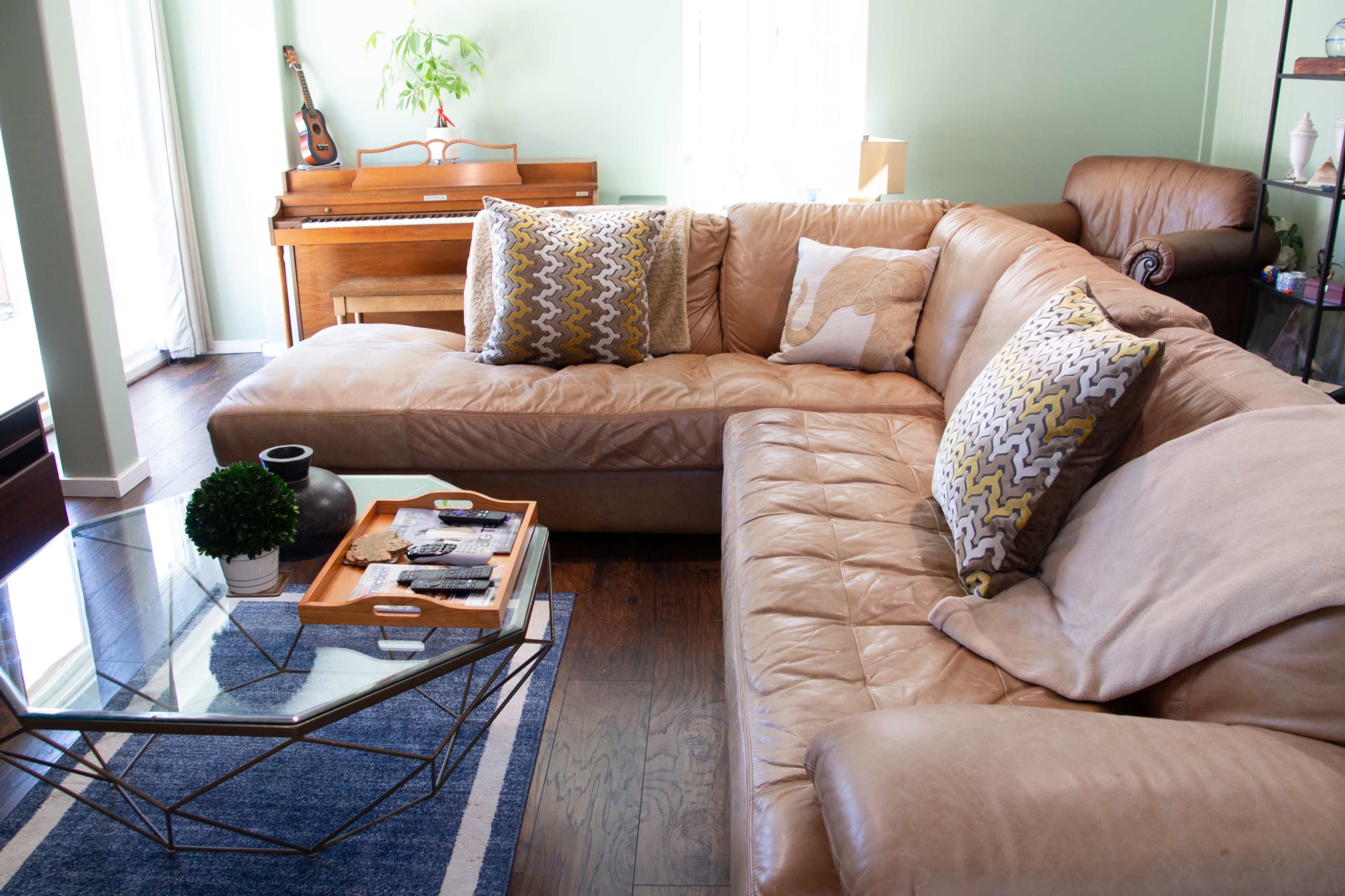 The living room features a brown leather sectional sofa with patterned cushions, a glass coffee table with a tray and magazines, and a wooden piano in the background.