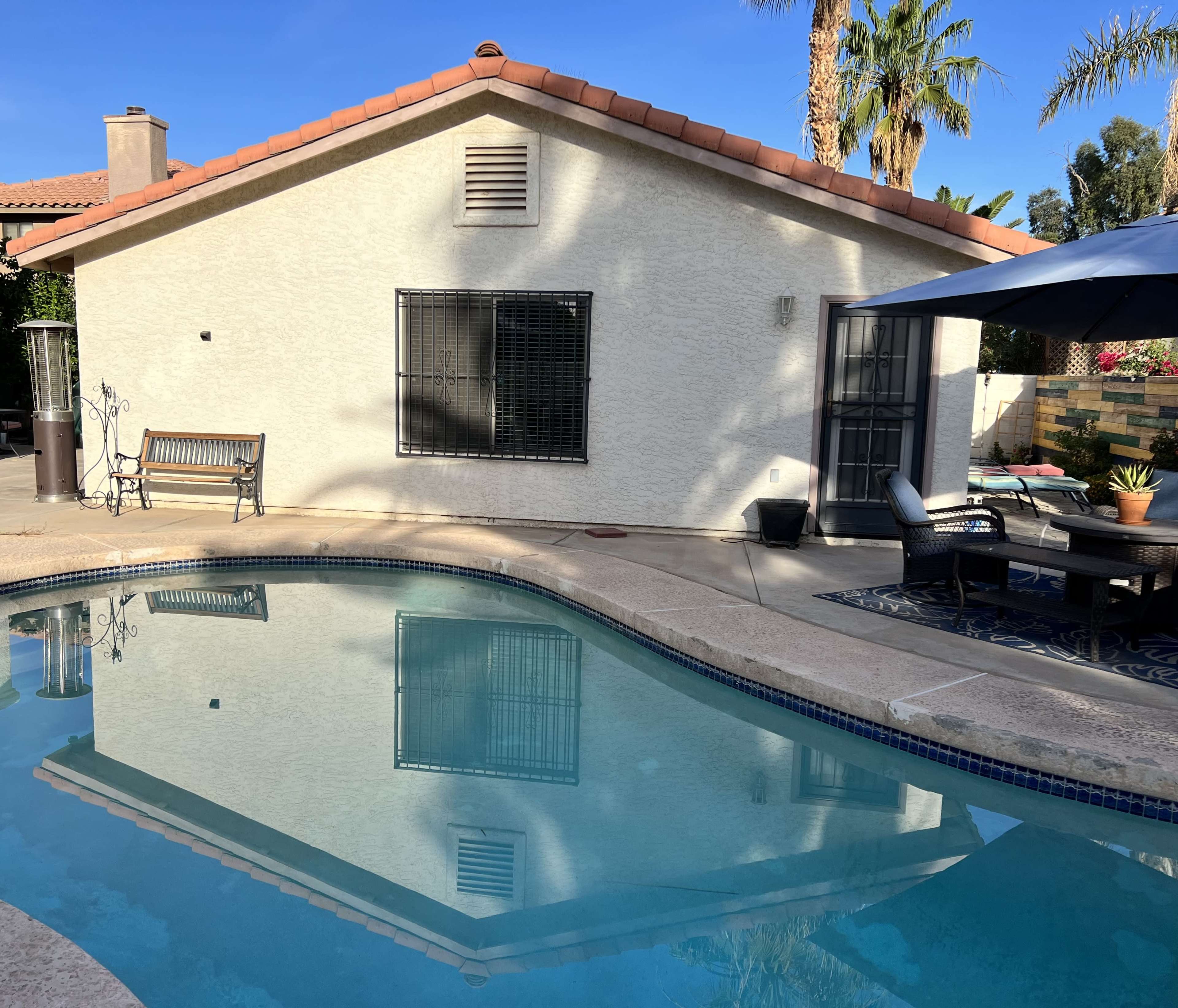A swimming pool reflecting a house with a patio and seating area adjacent to it, surrounded by palm trees.