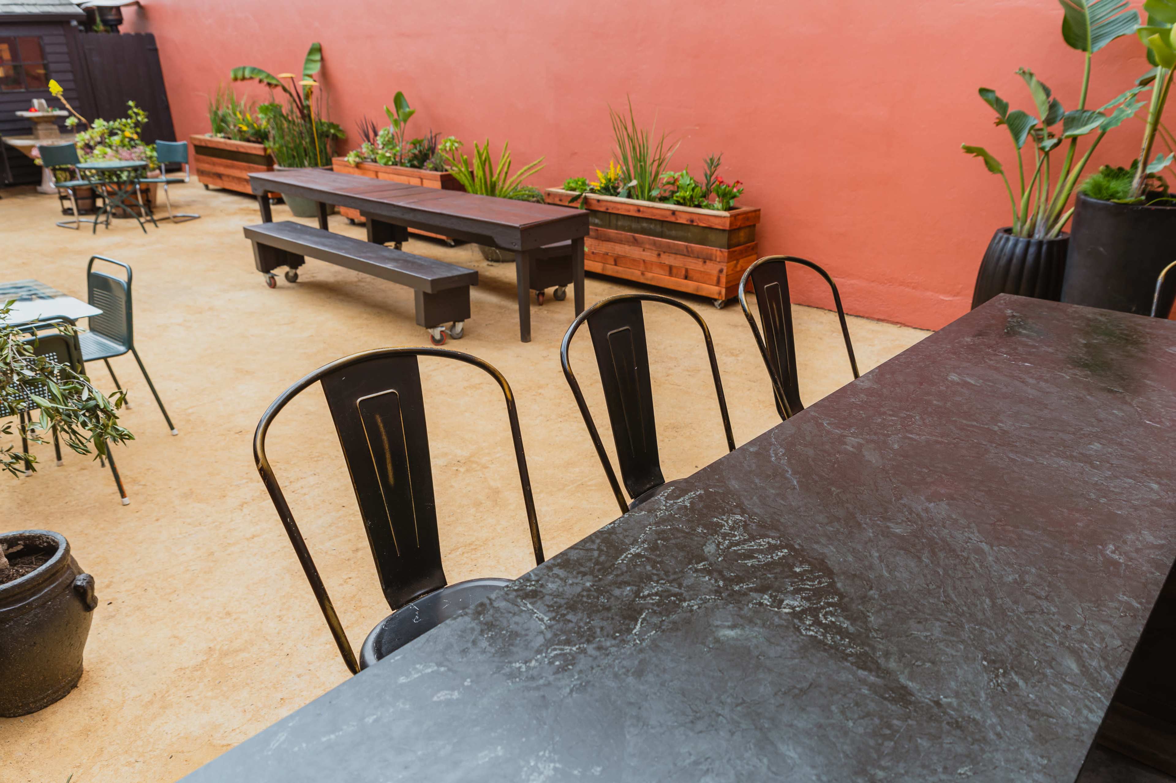 An outdoor seating area with black chairs and tables, surrounded by potted plants against a red wall.