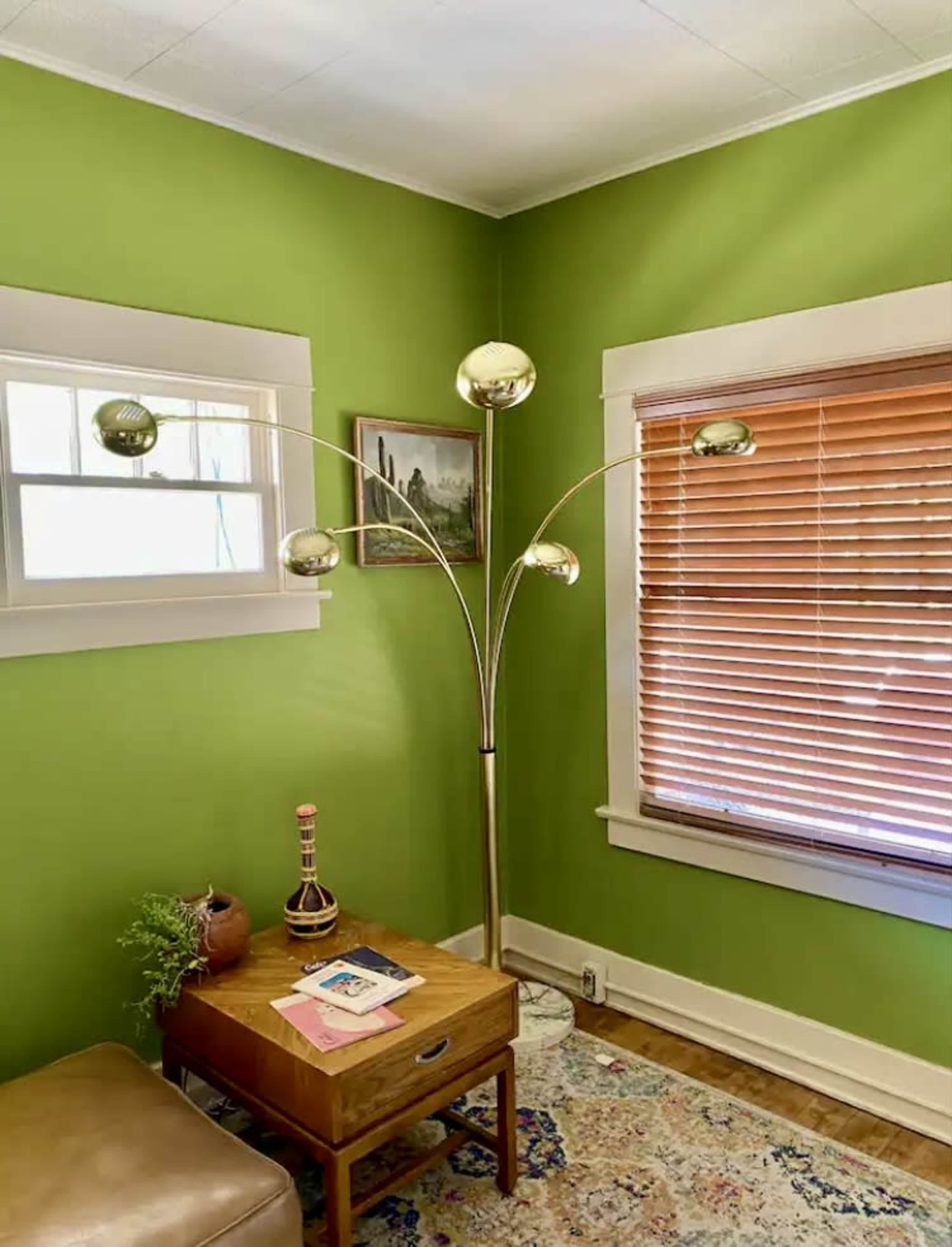 A floor lamp with four spherical lights stands next to a wooden table in a green-walled room with blinds on the window.