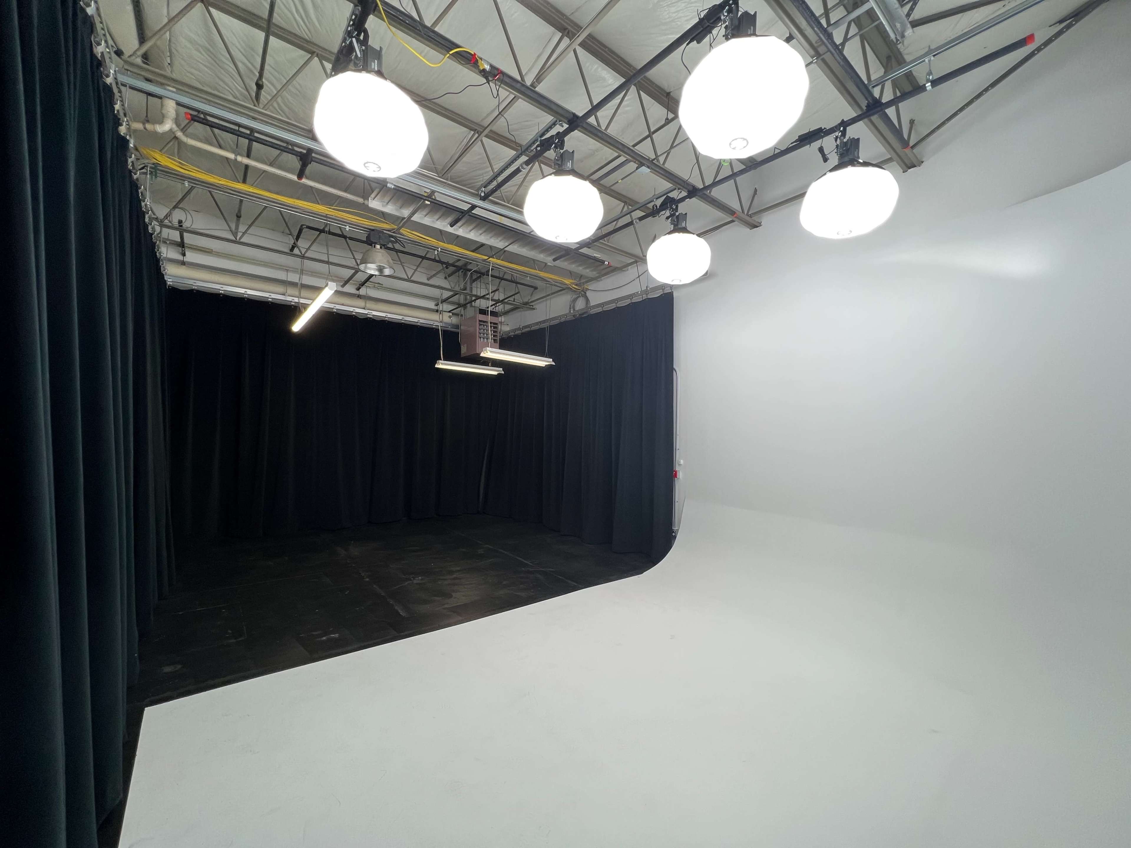A spacious photography studio with a white seamless backdrop, surrounded by black curtains and overhead lighting fixtures.