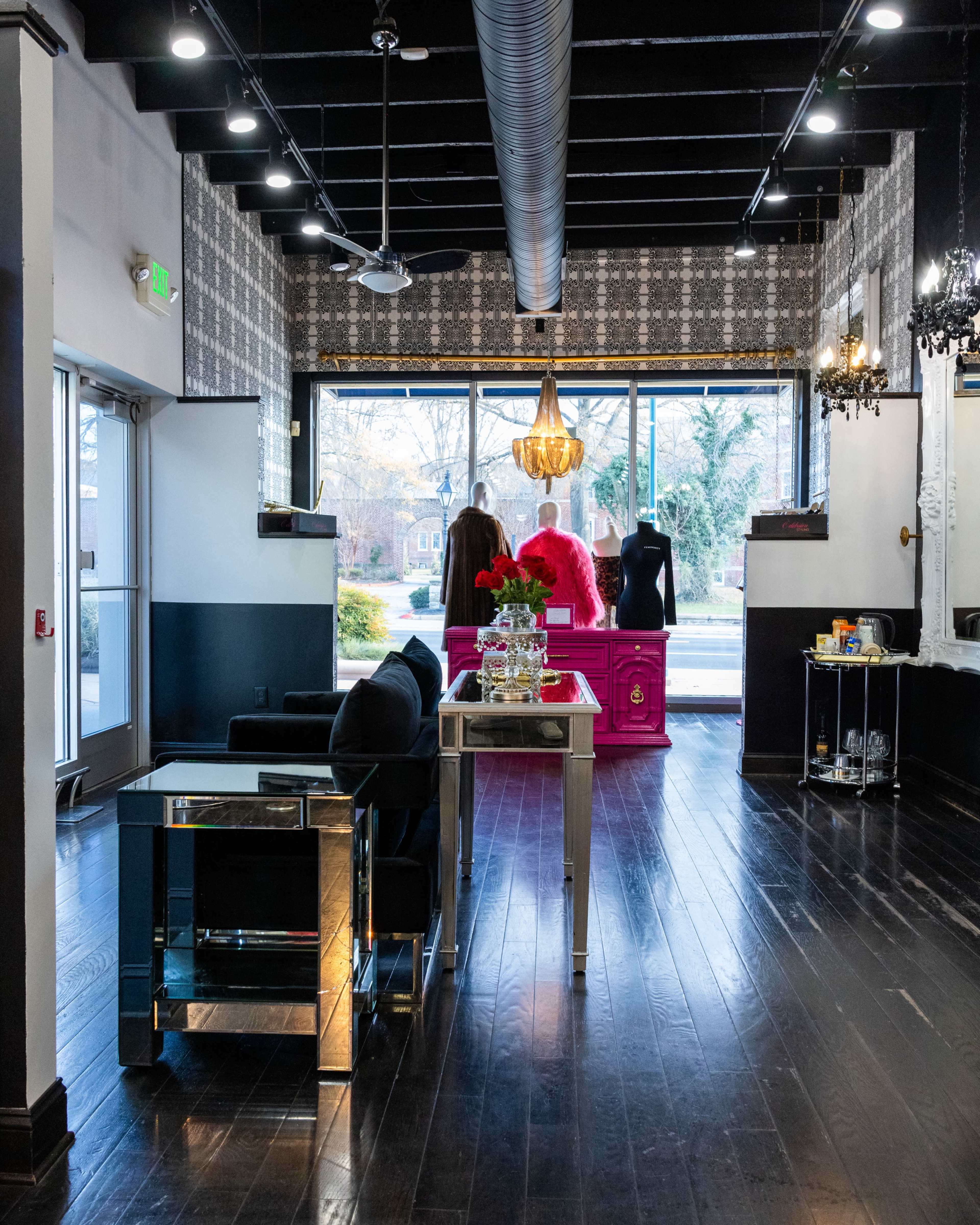 The image shows a modern boutique interior featuring a reception area with a glass table, black chairs, and a brightly colored pink display in front of large windows.