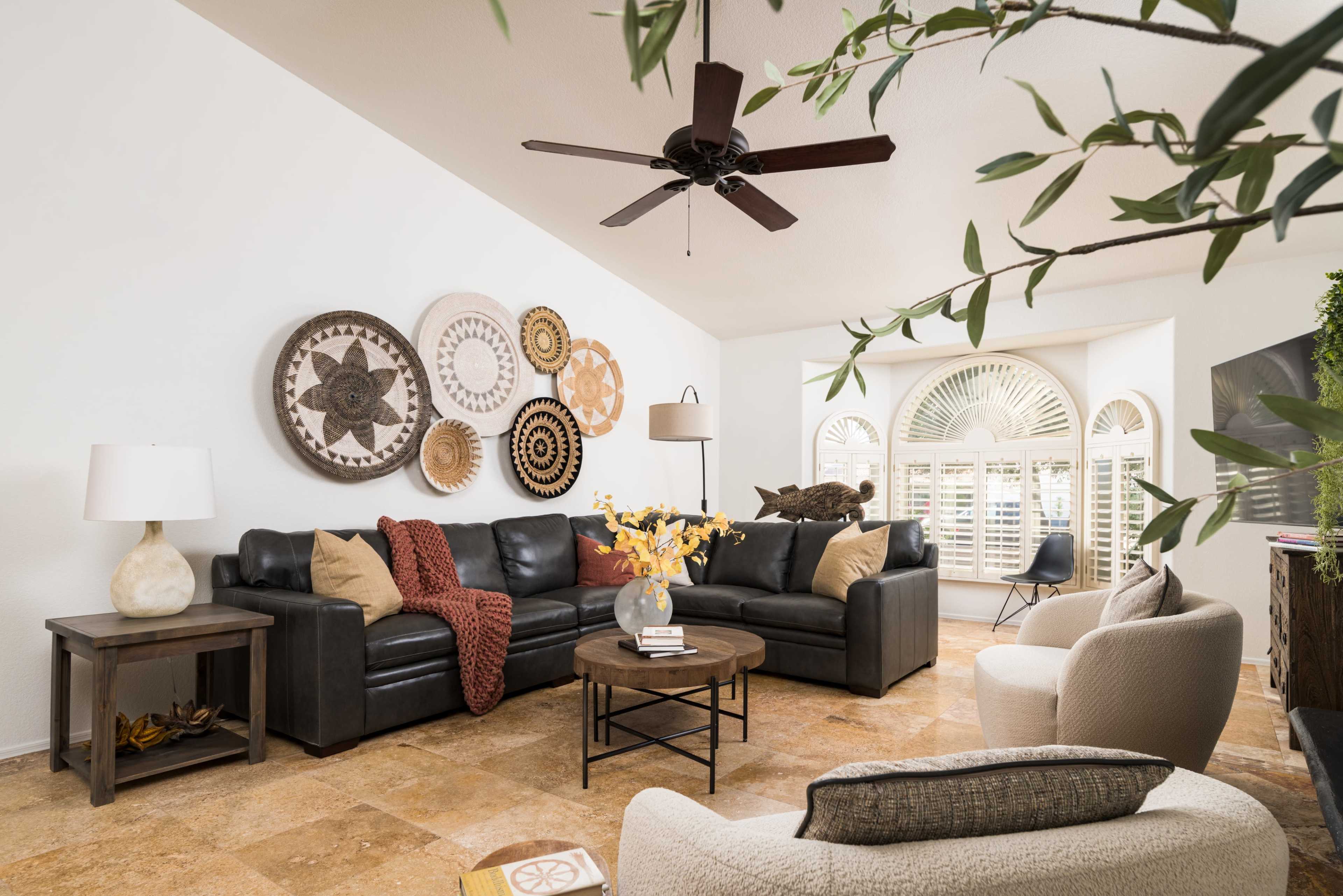 The living room features a black leather sectional sofa, a round coffee table, and decorative wall art, illuminated by natural light from large windows.