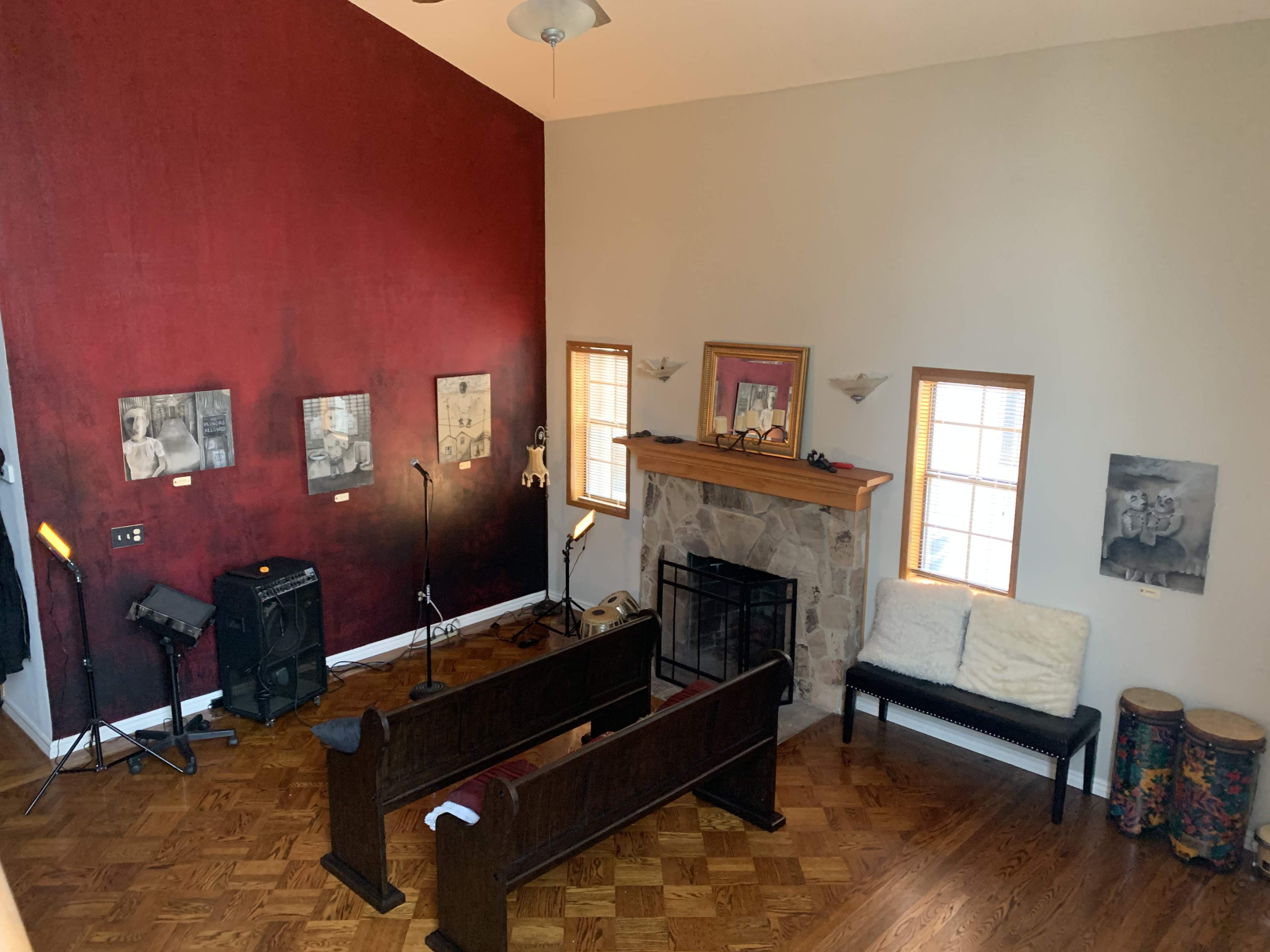 The image shows a spacious living room with a high ceiling, featuring a red accent wall, a stone fireplace, wooden benches, and various pieces of artwork displayed on the walls.