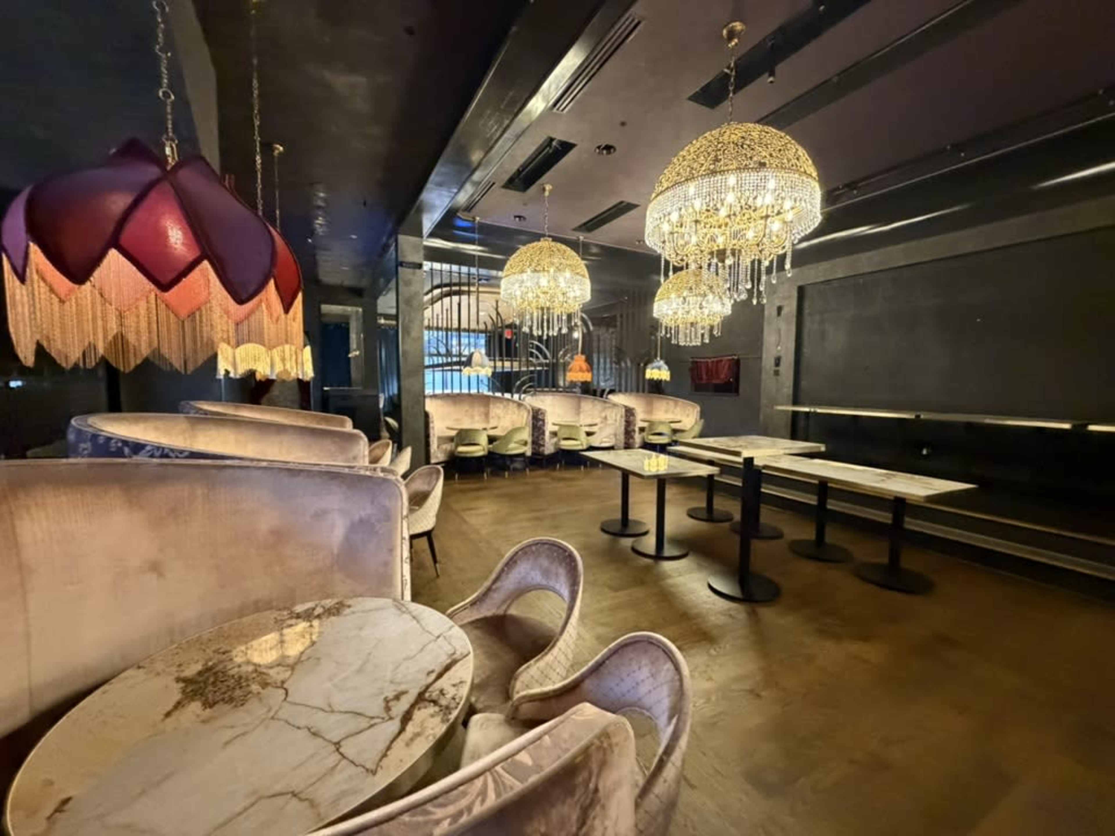 The image shows a dimly lit lounge with plush seating, ornate chandeliers, and marble tables.