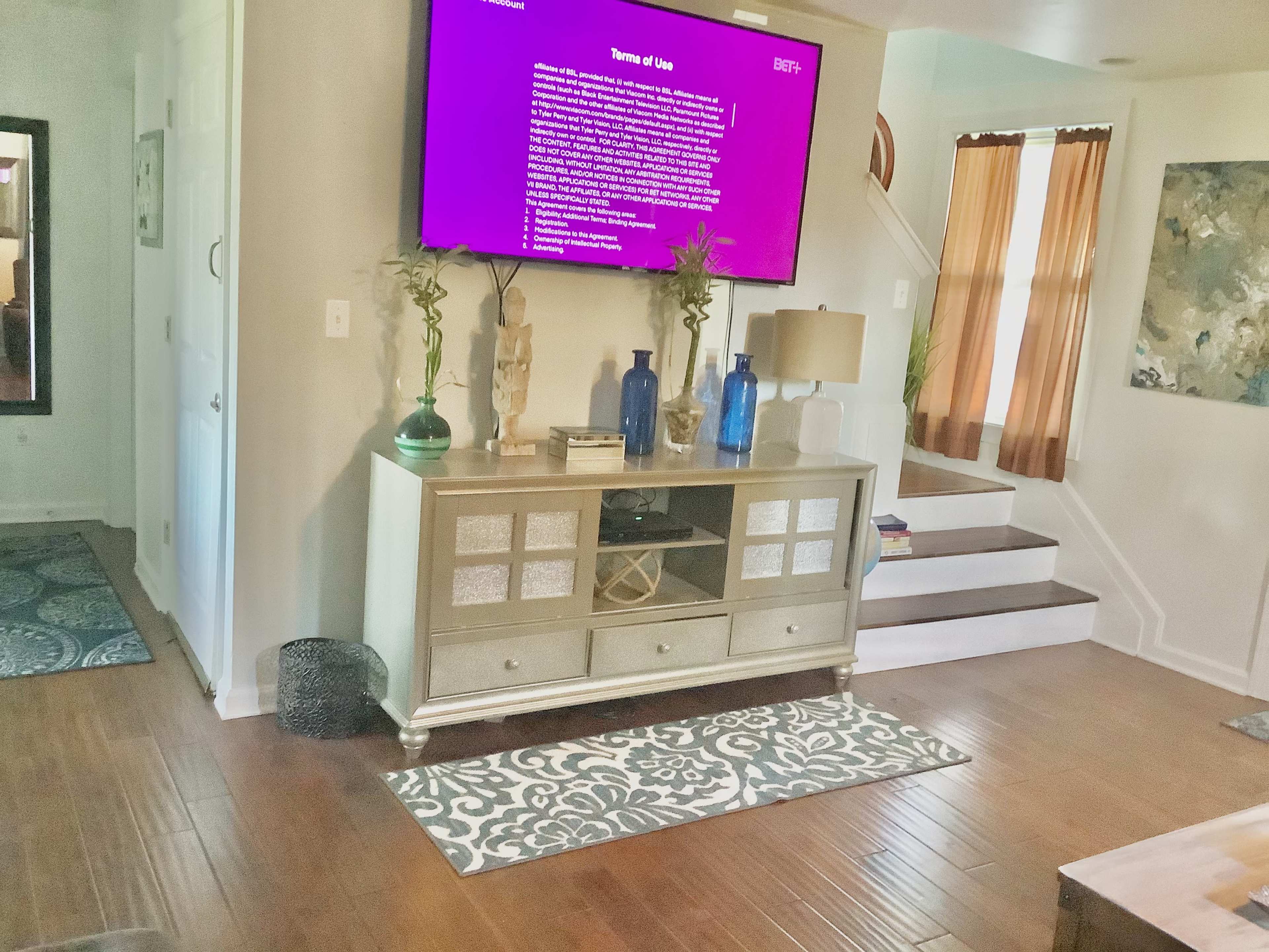 The room features a modern entertainment center with a large TV mounted above it, surrounded by decorative vases and a staircase leading to an upper level, with two windows draped in sheer curtains.