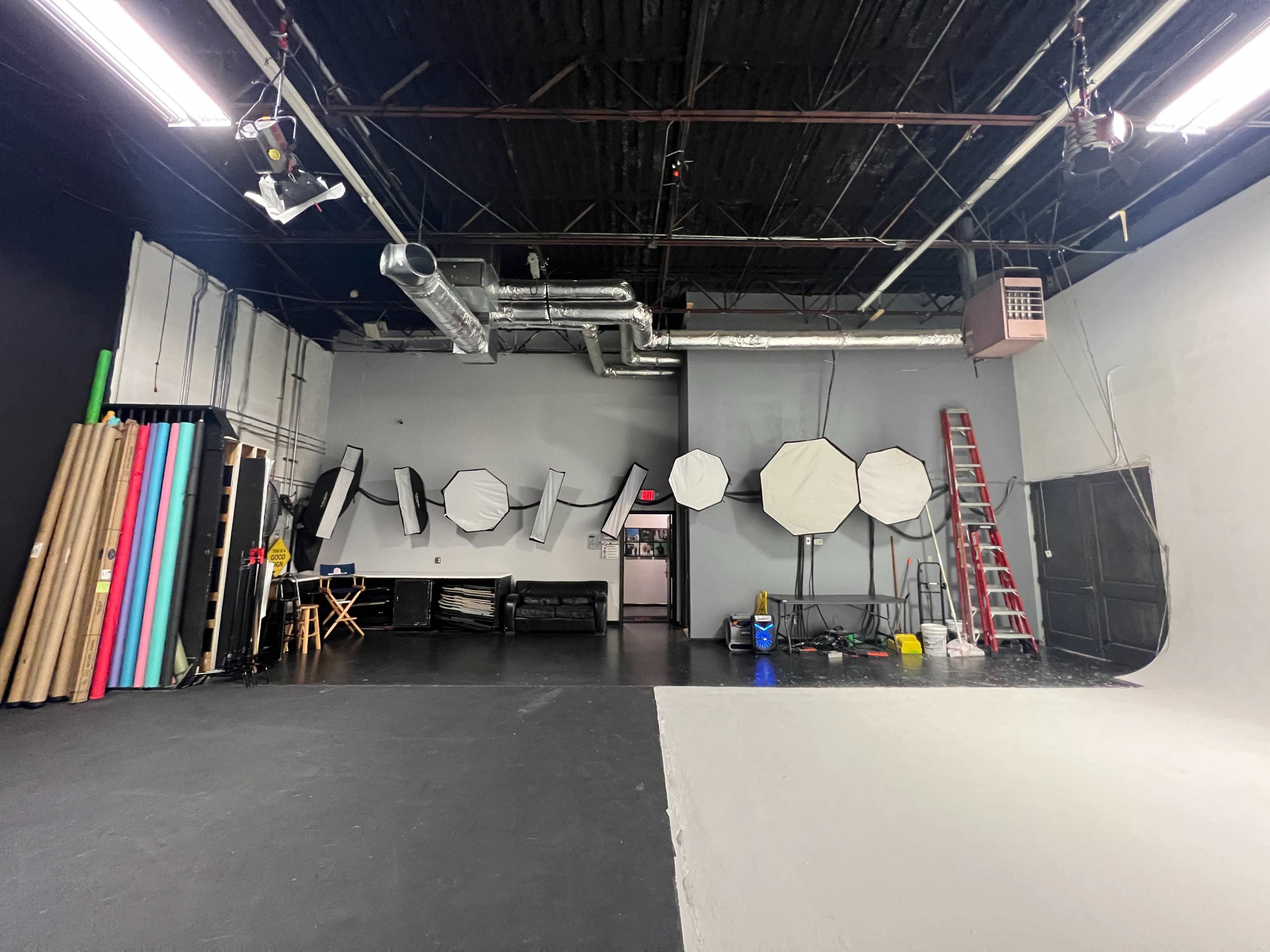 The image shows a photography studio with a black wall, various backdrops, lighting equipment, a ladder, and a seating area.