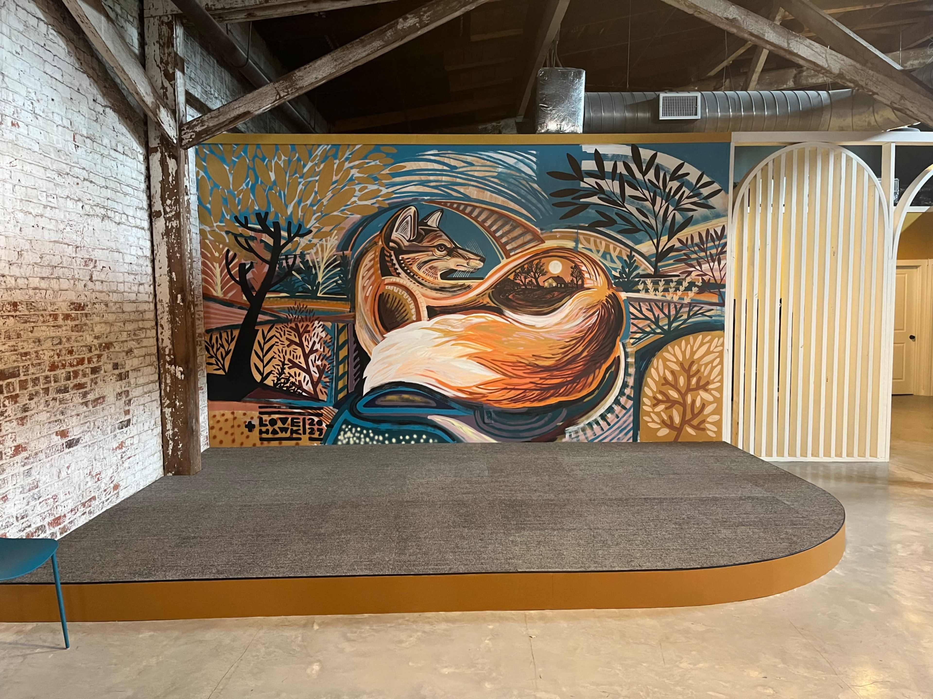 The image shows a vibrant mural of a fox among stylized trees on a bold-colored wall, with a raised platform in front.