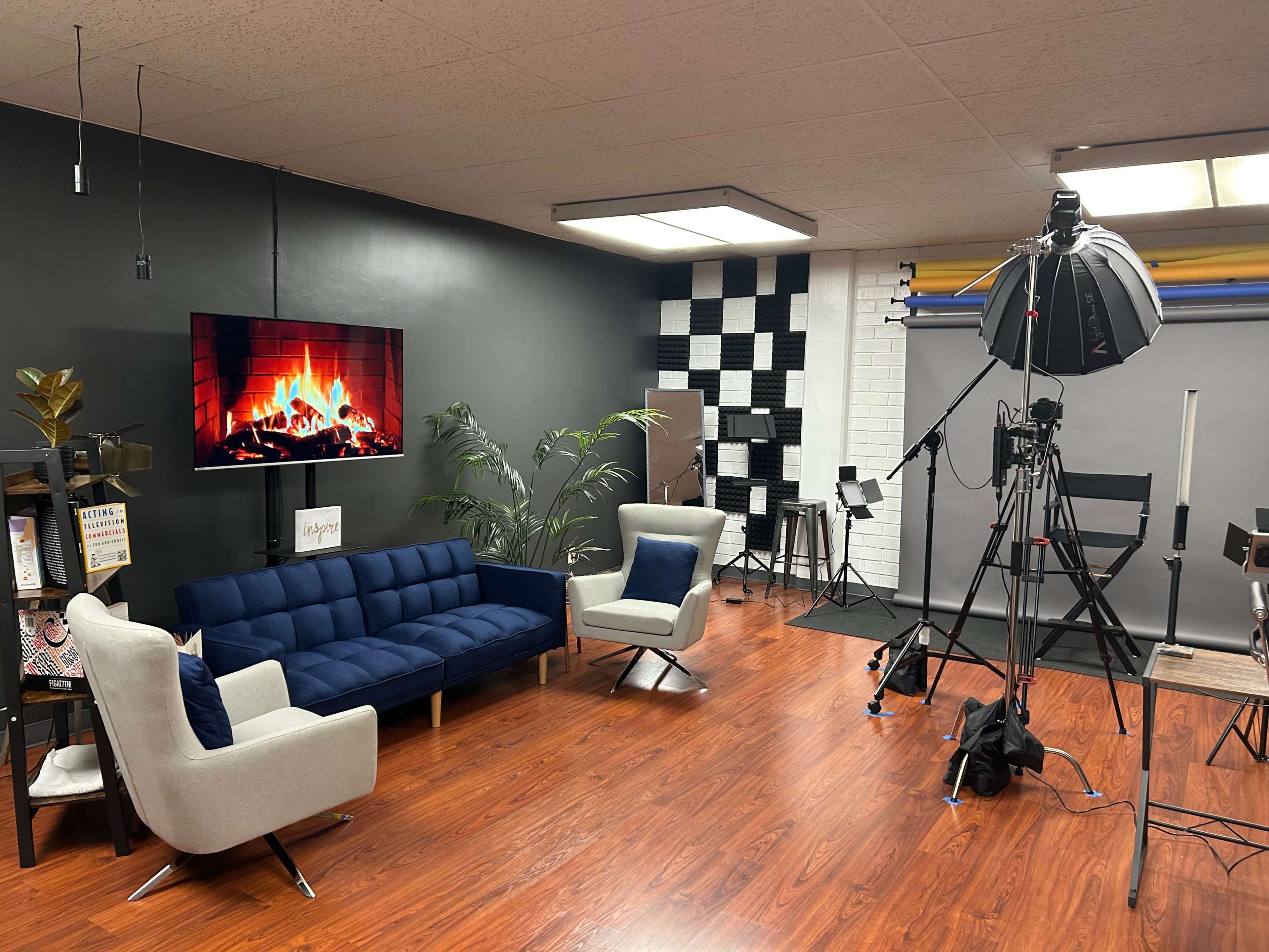 A modern studio setup featuring a blue couch, an artificial fireplace display on the wall, and various filming equipment including lighting and tripods.