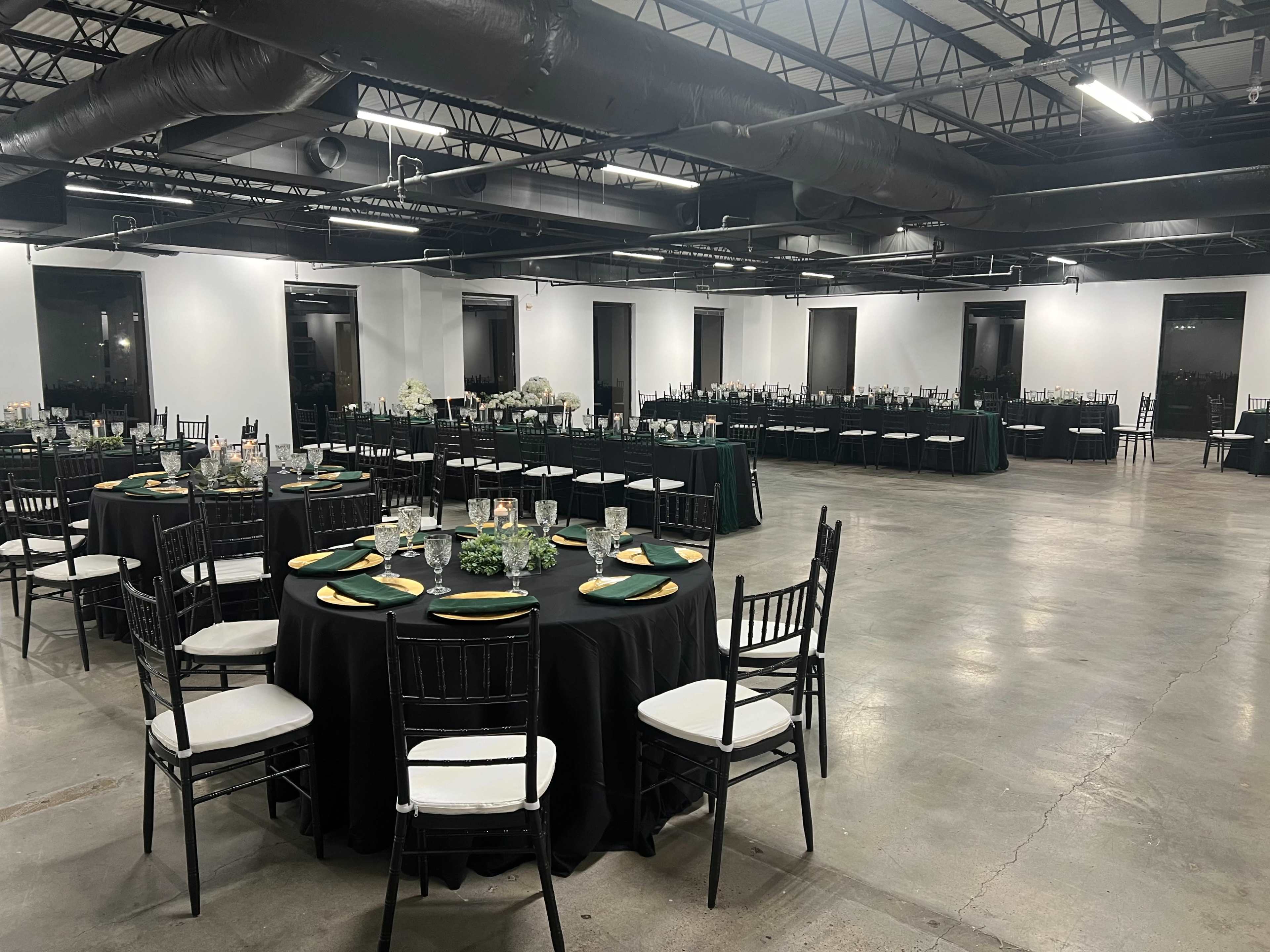 A spacious banquet hall is set with black tablecloths, round tables arranged with dishes and glassware, and green accents throughout.