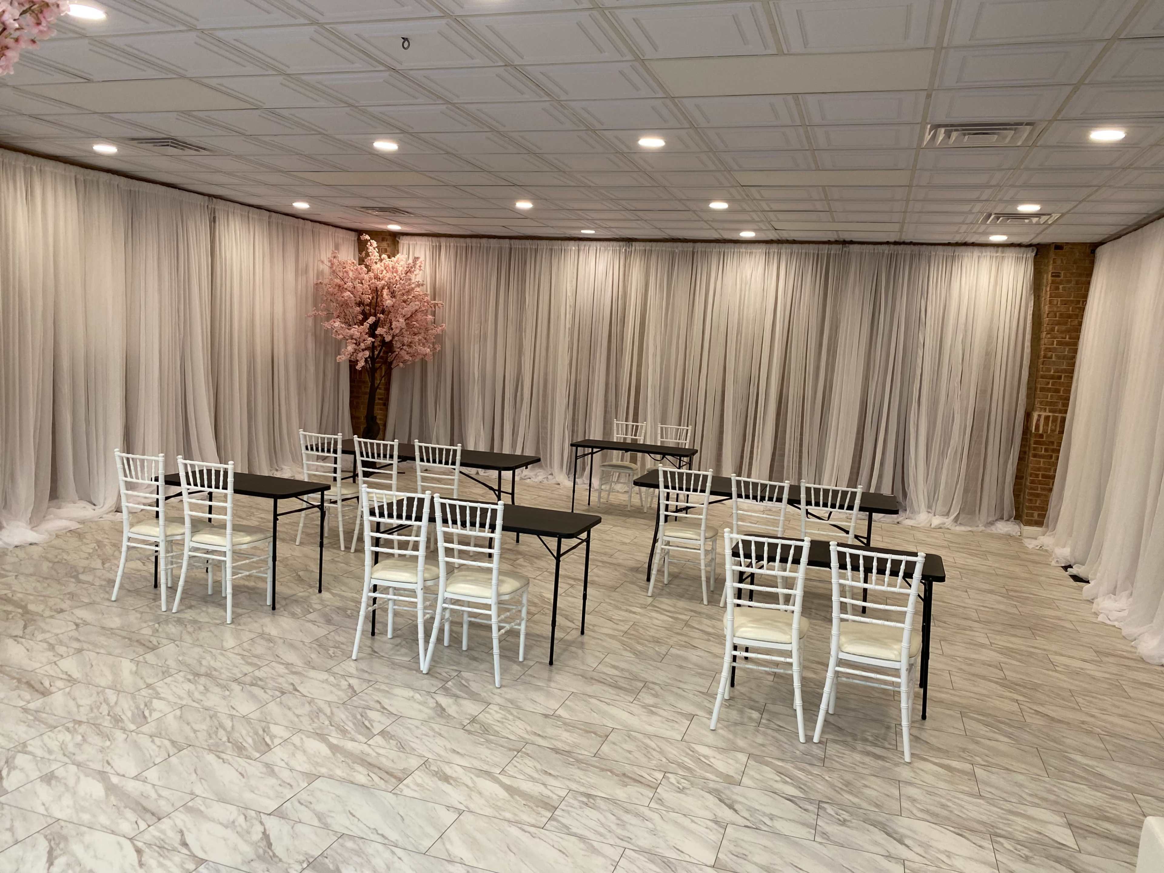 A room with white draped curtains, several folding tables arranged in a U-shape, and white chairs set around them, with a pink cherry blossom tree in the corner.