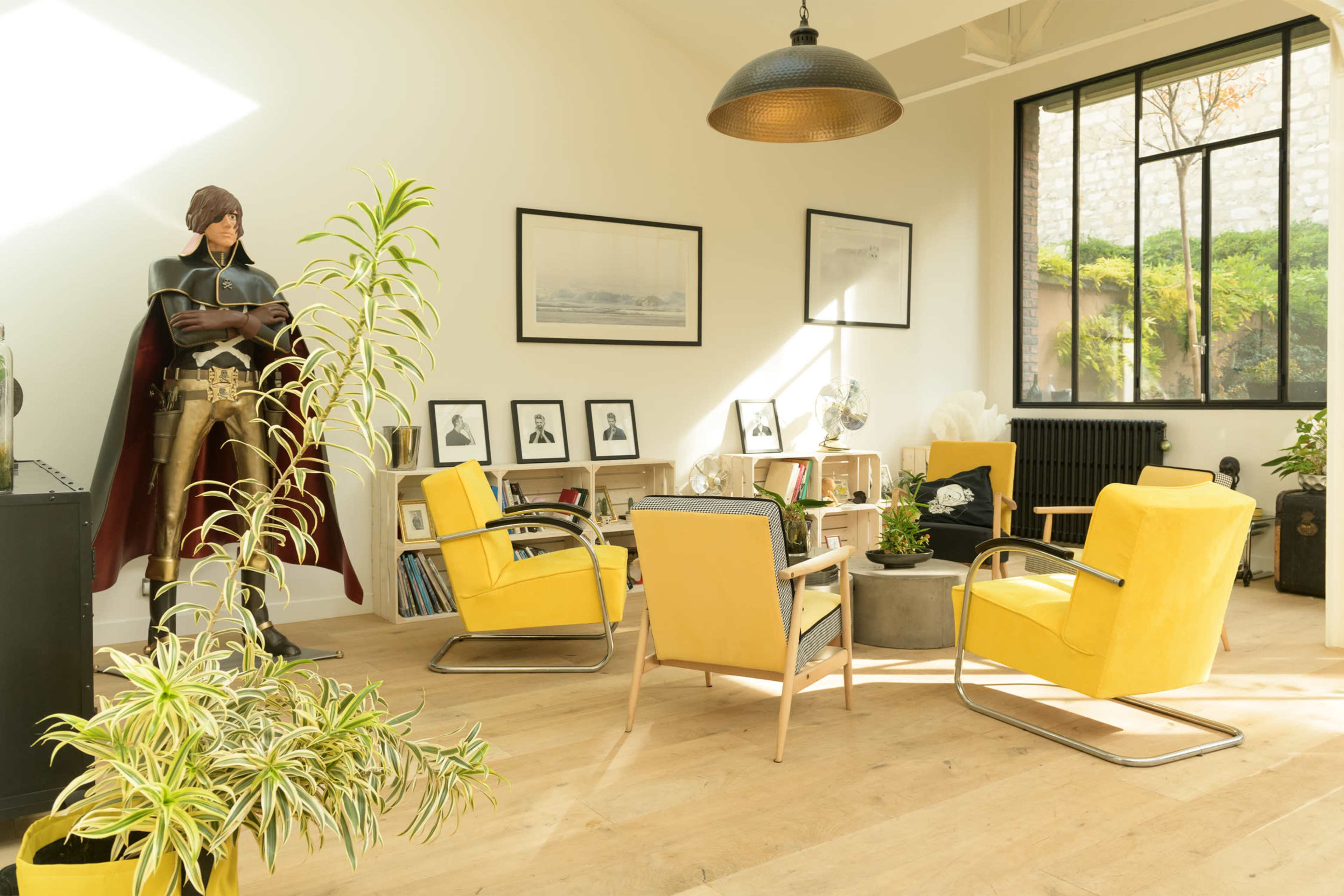 A modern living room with yellow chairs, a large plant, a bookshelf, and a life-sized figure in a superhero costume standing in the corner.