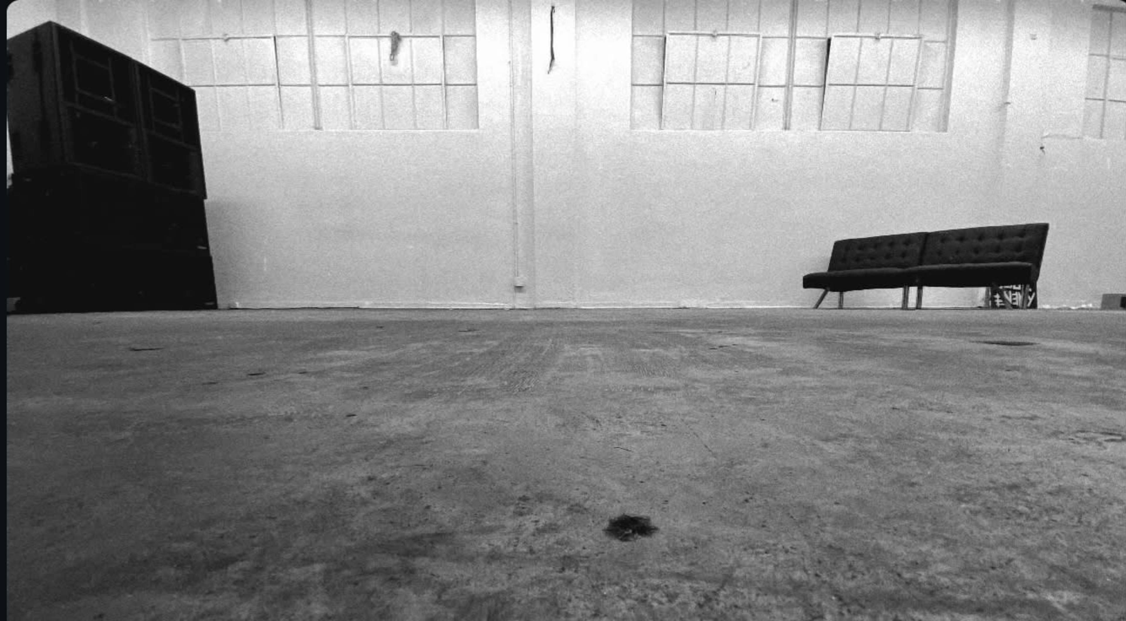 The image shows an empty, concrete-floored interior with large windows, a black speaker stack in the corner, and a modern couch against the wall.