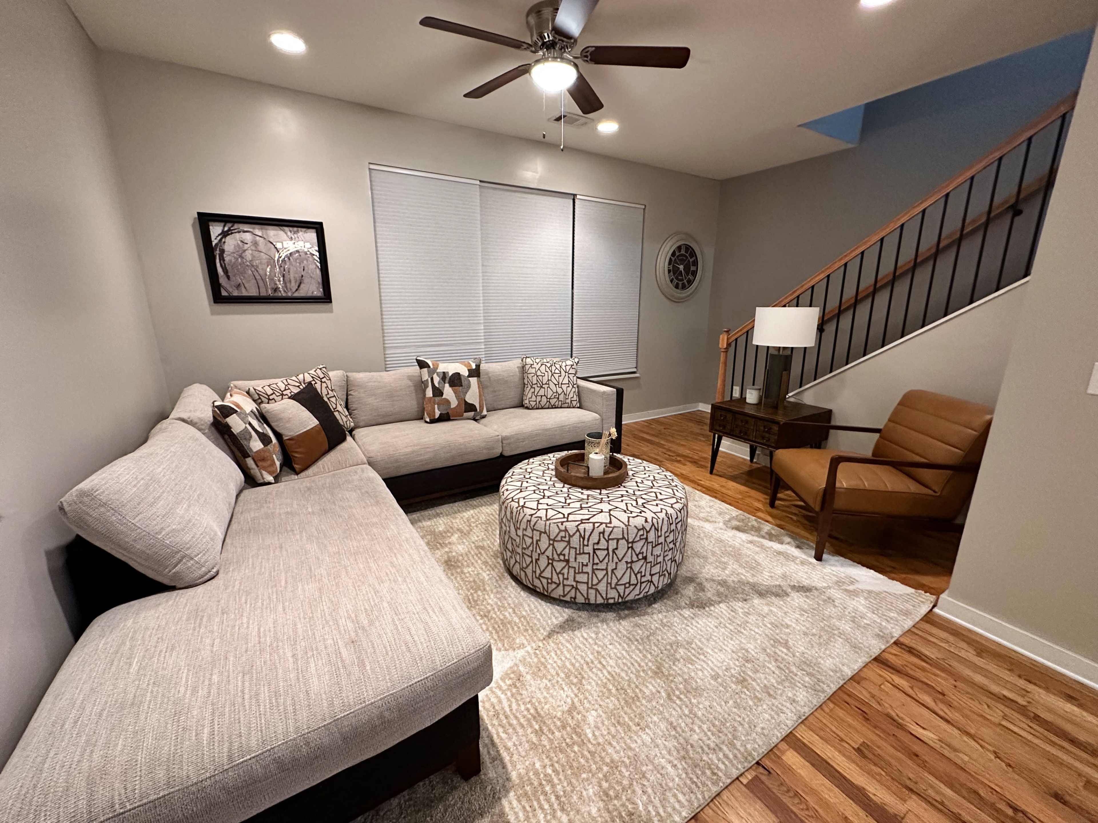 A modern living room featuring a sectional sofa, a circular coffee table, an armchair, and large windows with blinds.