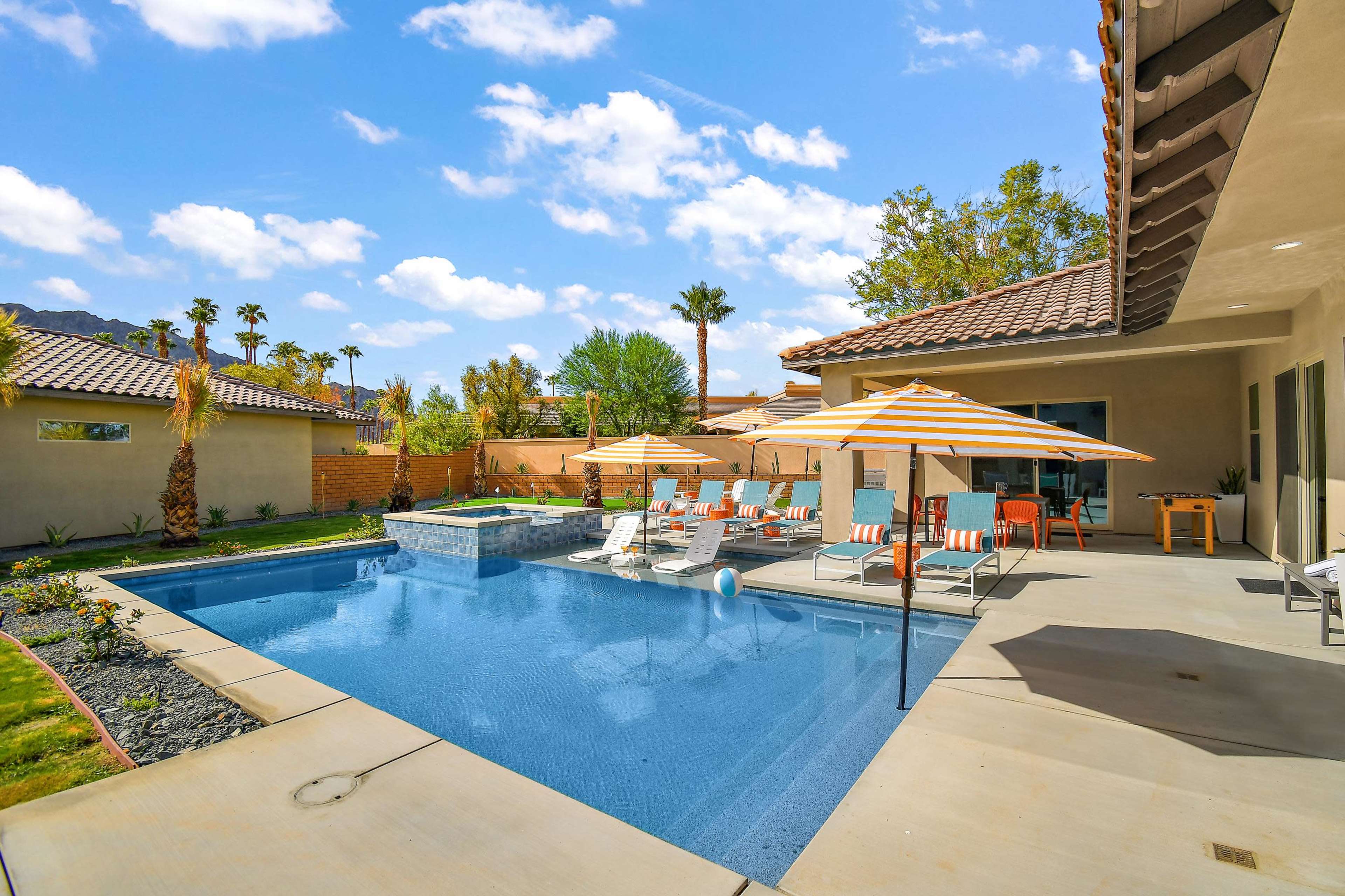 A residential backyard featuring a swimming pool, several lounge chairs with umbrellas, and landscaped greenery under a blue sky.
