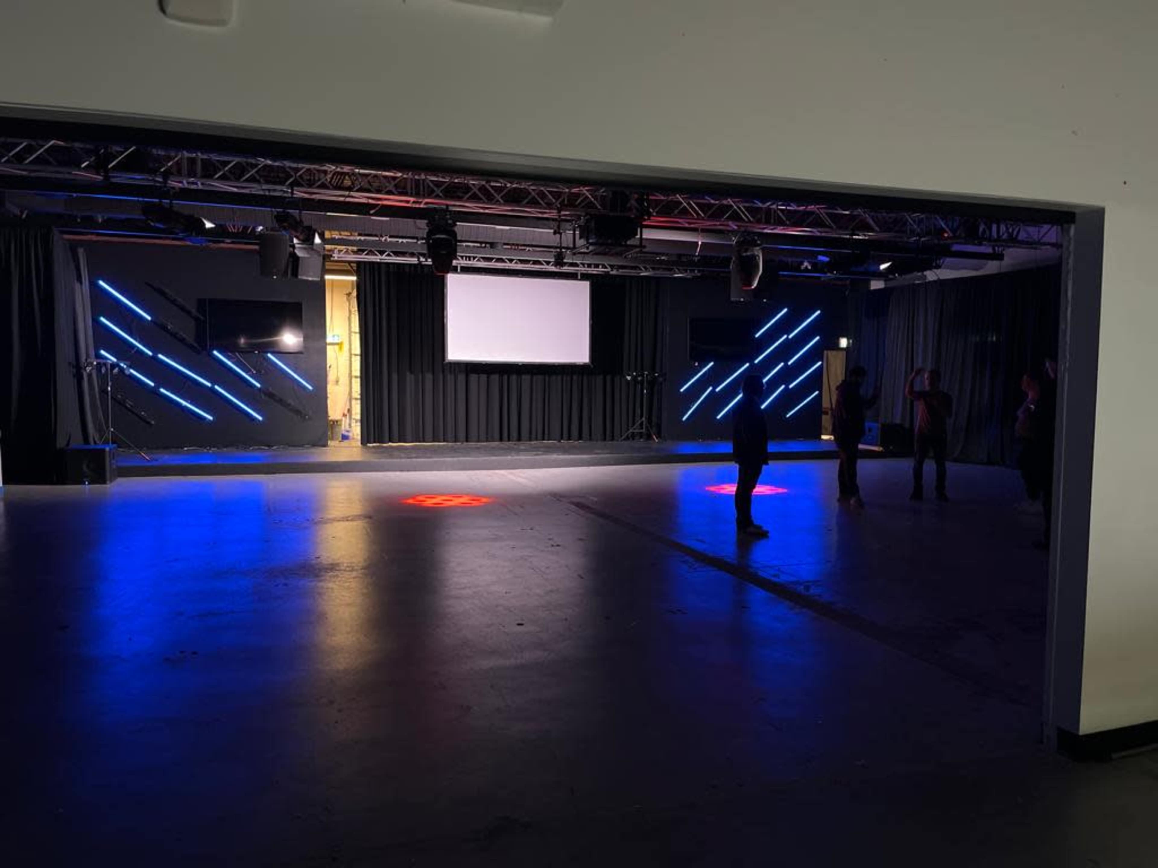 A dimly lit, empty performance space with colorful LED lights and a projector screen at the front.