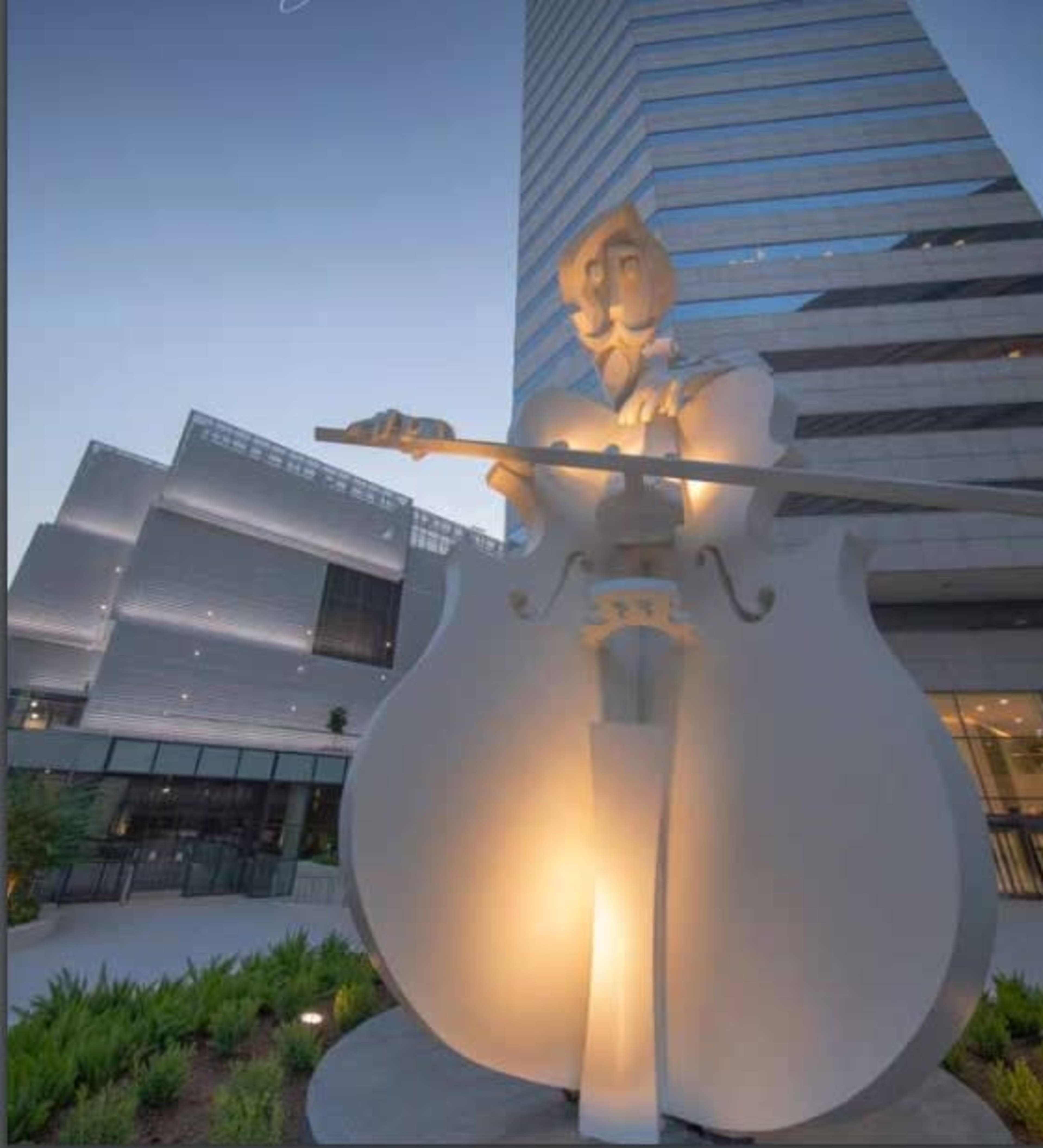 A large, illuminated statue of a musician playing a cello stands in front of a modern building.