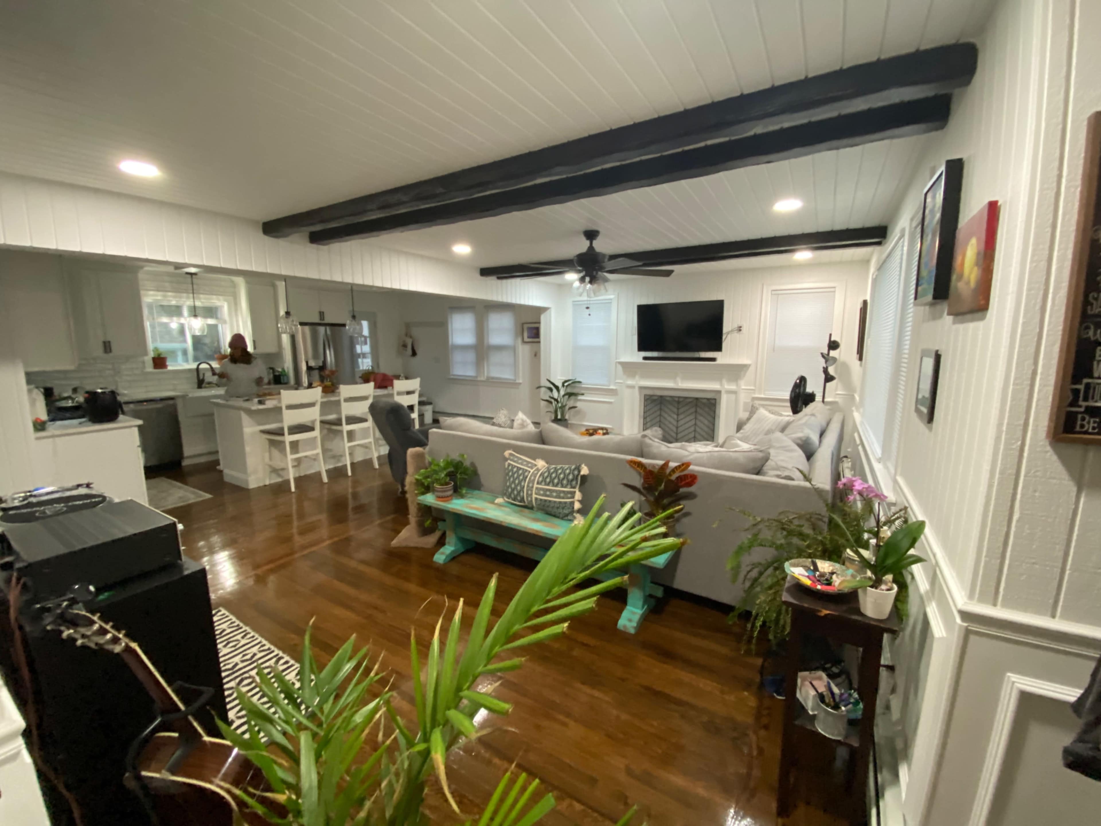 The image shows a living room with a grey sofa, wooden beams on the ceiling, a coffee table, and a view of a kitchen area with white furniture in the background.