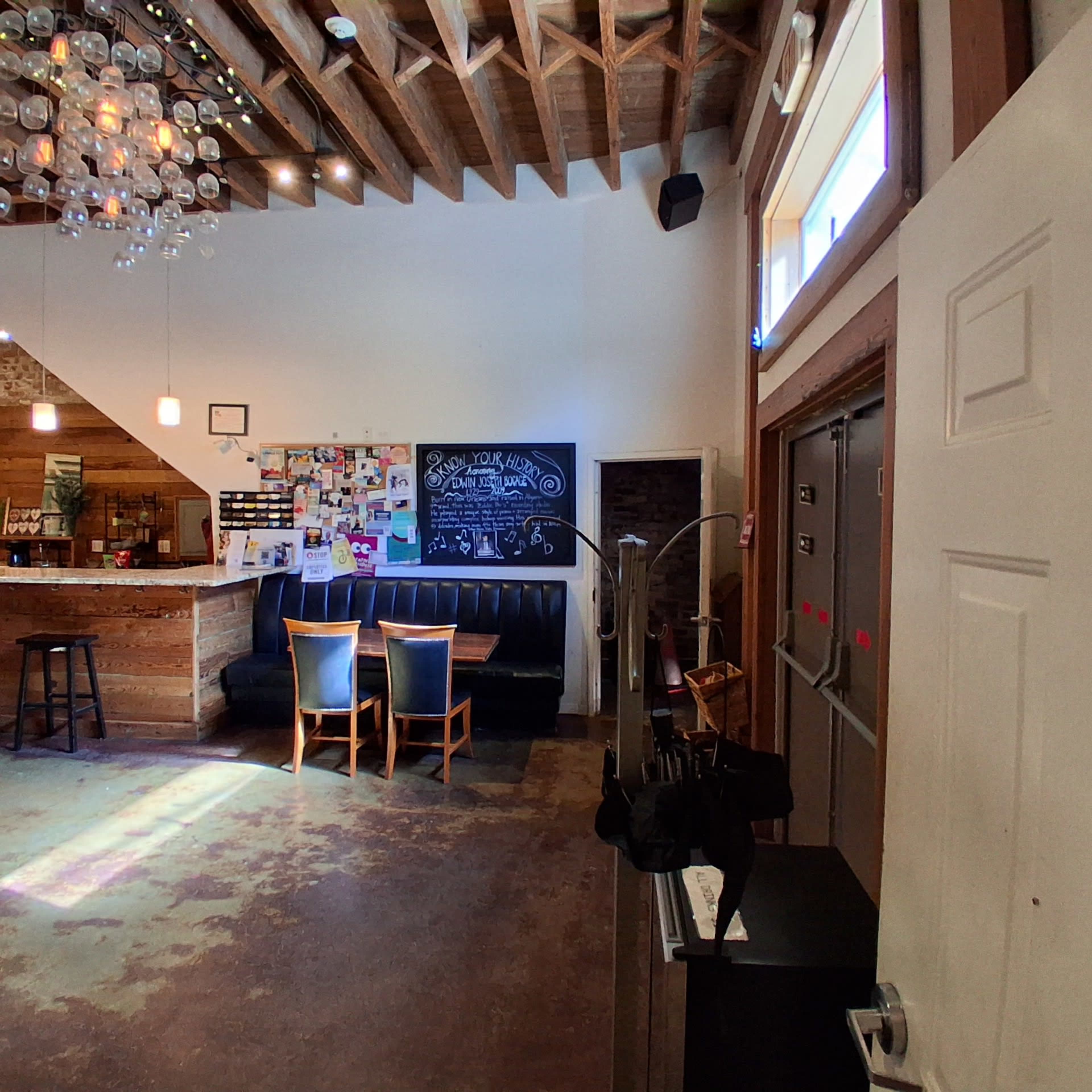 The image shows a spacious interior of a café or restaurant, featuring a seating area with black cushioned chairs, a chalkboard wall, and a bar counter with pendant lighting above.