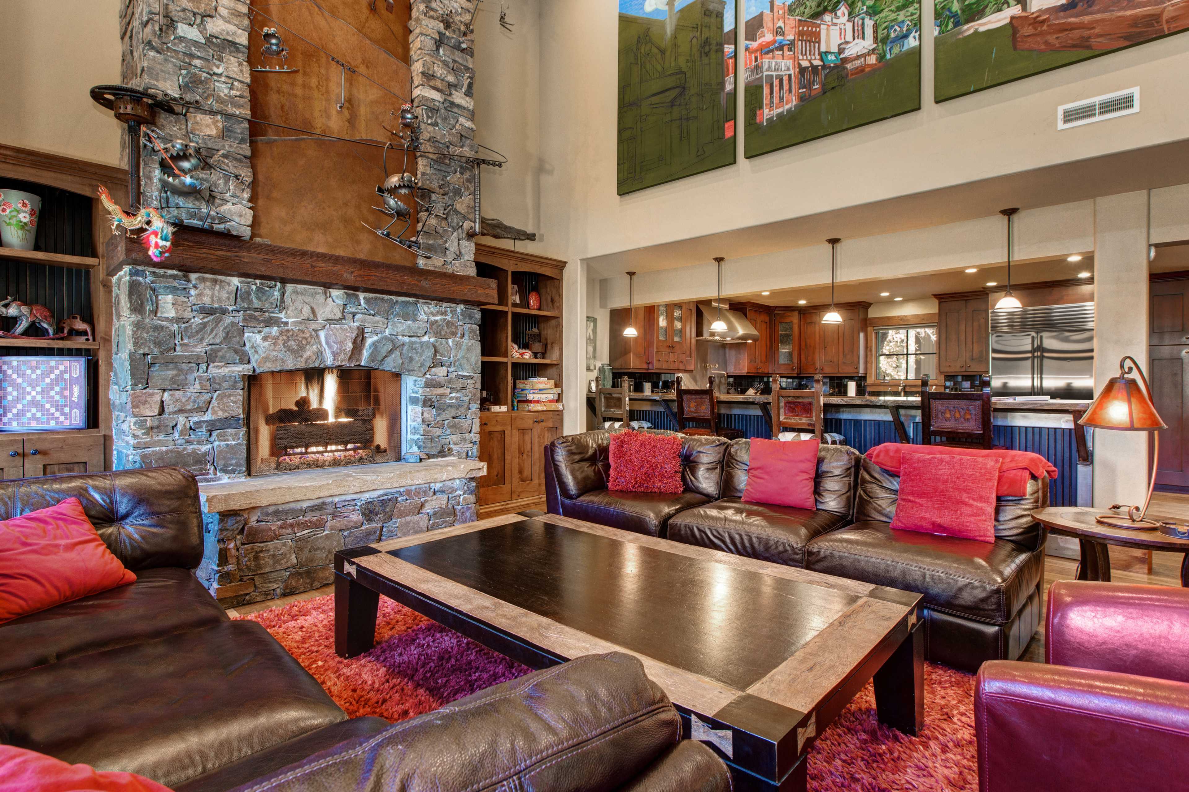 A spacious living room with a stone fireplace, dark leather sofas, a large coffee table, and an open kitchen in the background.