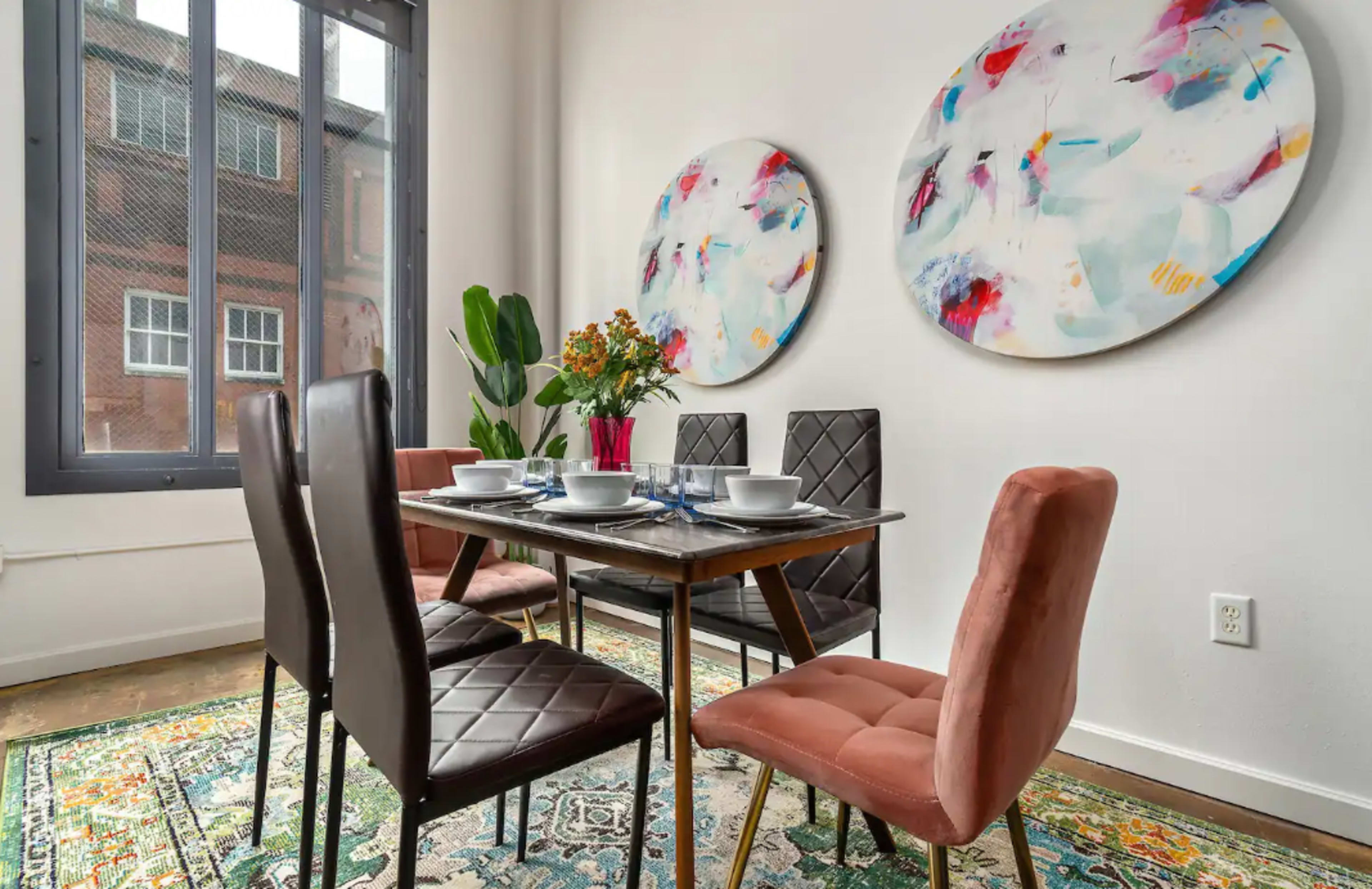 A dining area features a table set for four with colorful wall art and a patterned rug.