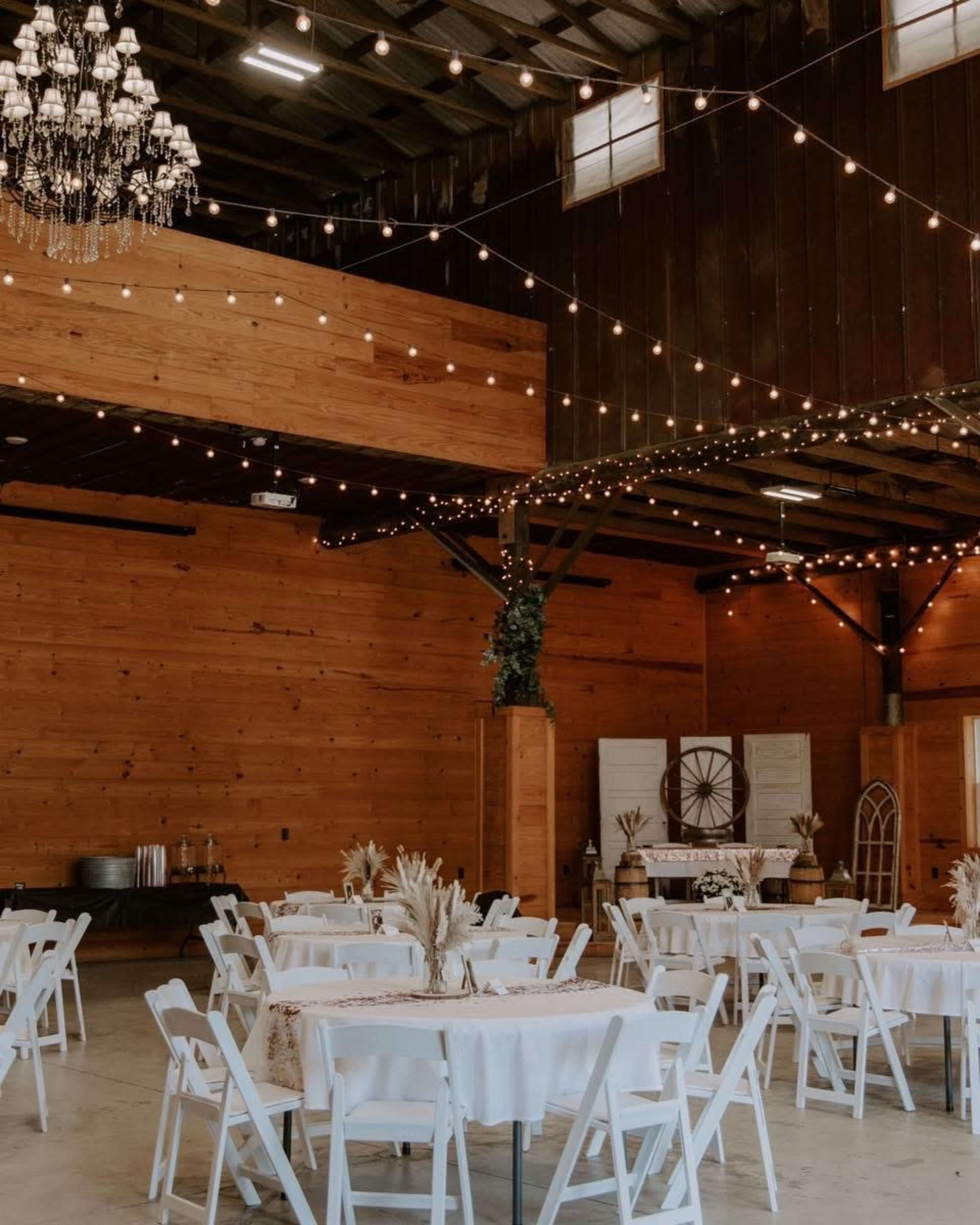 The image shows an interior banquet hall with wooden walls, round tables covered in white tablecloths, and string lights hanging from the ceiling.
