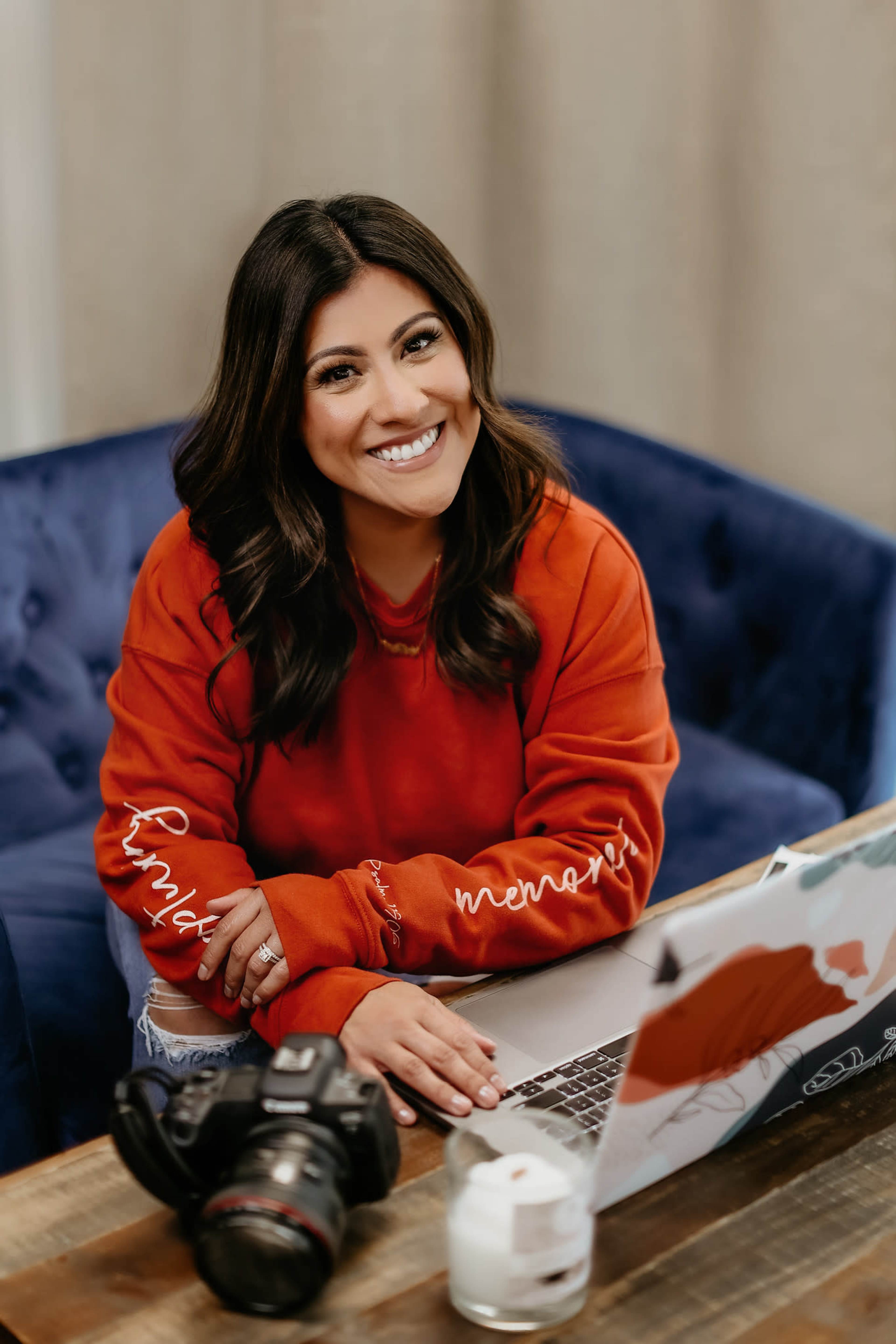 A woman in a red sweatshirt sits at a table with a camera and laptop in front of her, smiling at the camera.