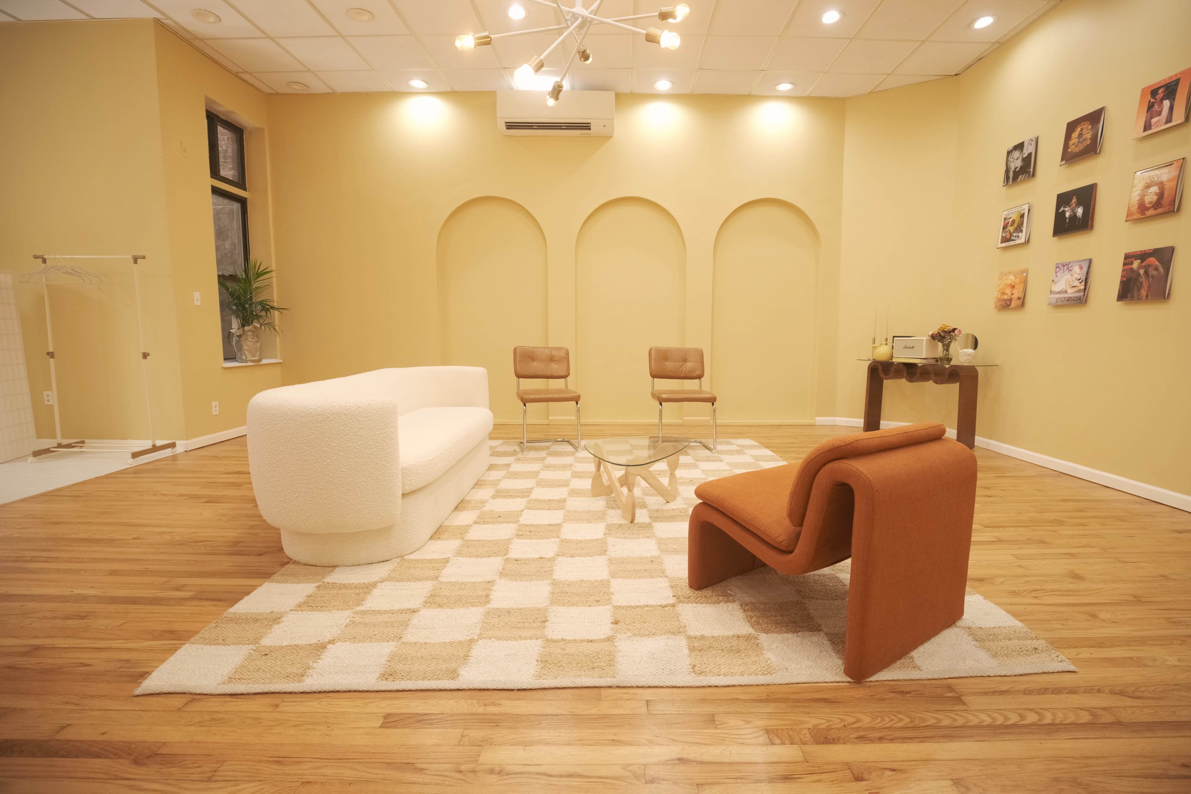 The scene features a modern interior with a curved white sofa, an orange chair, two tan chairs, a glass coffee table, and a checkered rug on a wooden floor, set against yellow walls with art displayed.