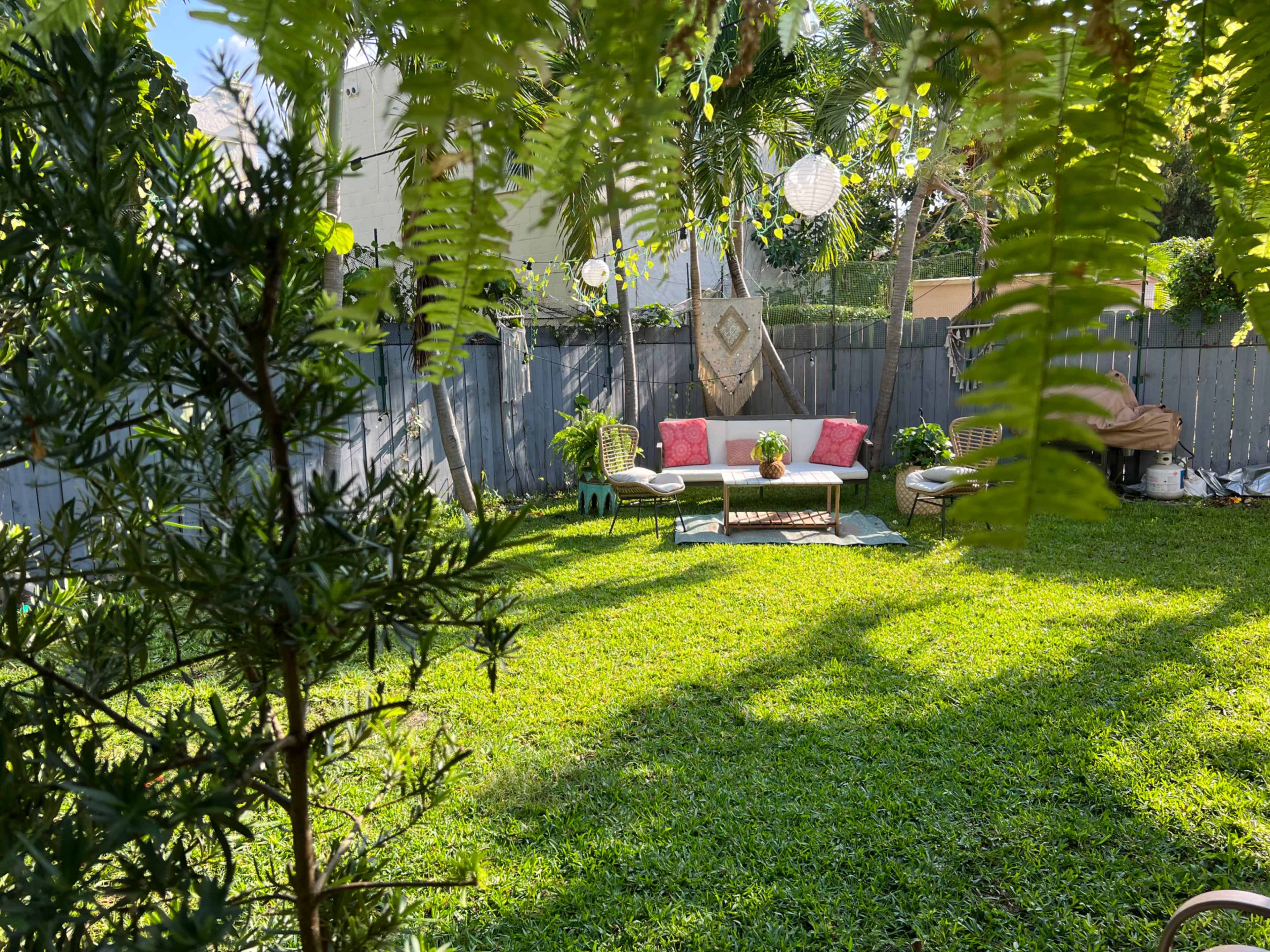 A cozy outdoor seating area with a table and pink cushions is set on a grassy lawn surrounded by plants and a wooden fence.