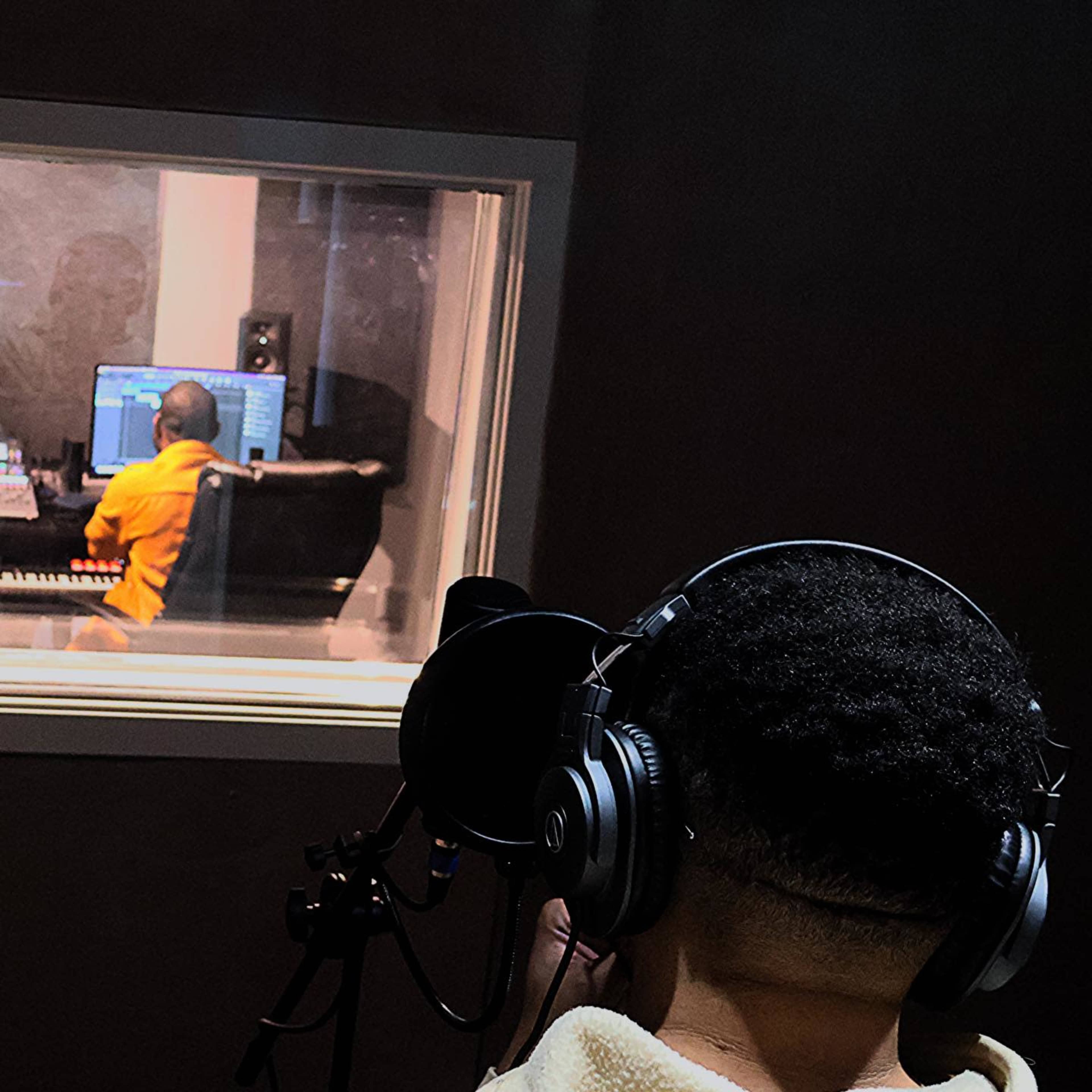 A person with headphones is recording vocals in a studio while another individual works on music production in a separate room.