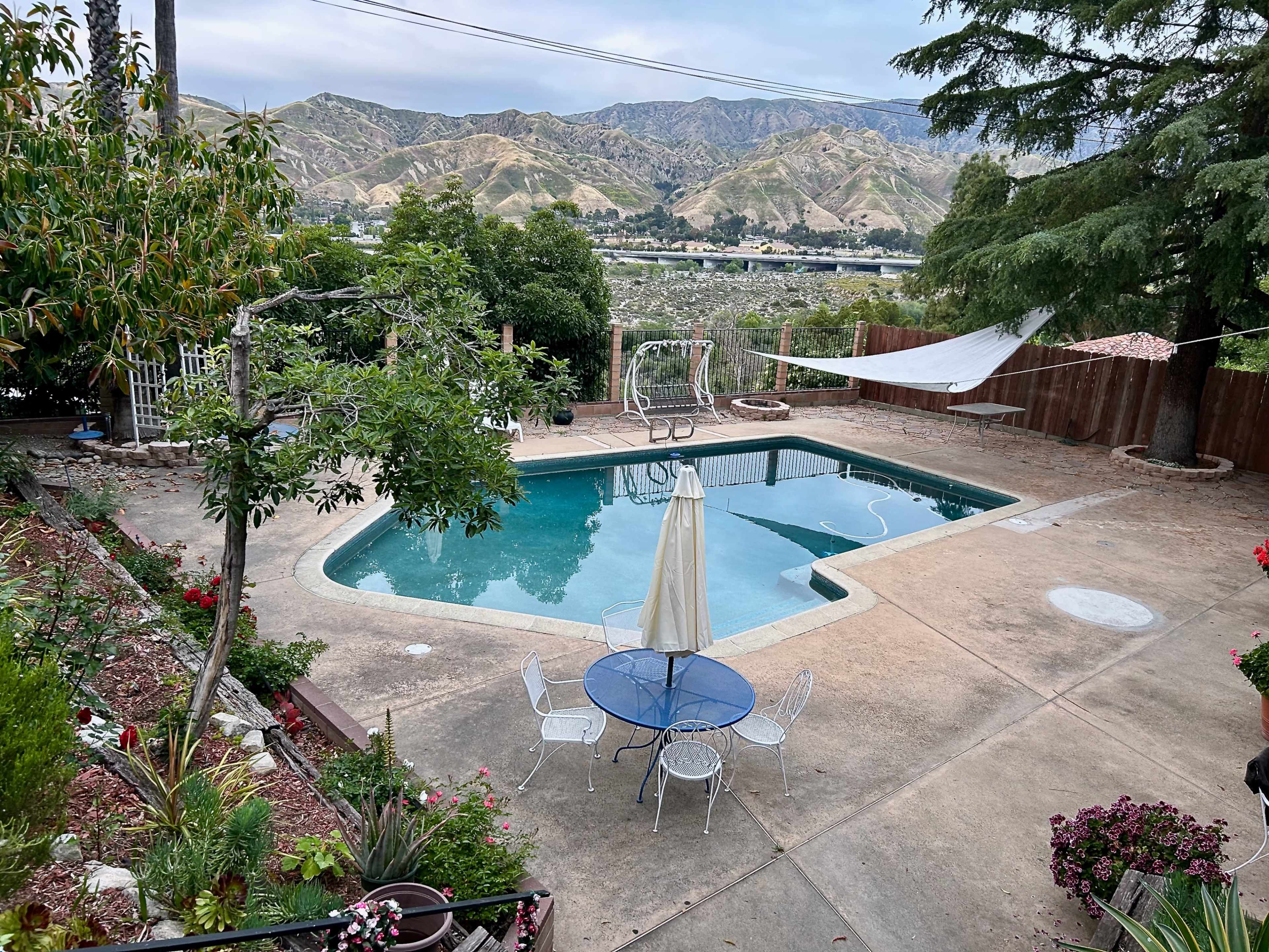 A backyard features a swimming pool surrounded by concrete, a garden area with flowers, a table with chairs, and distant mountains.