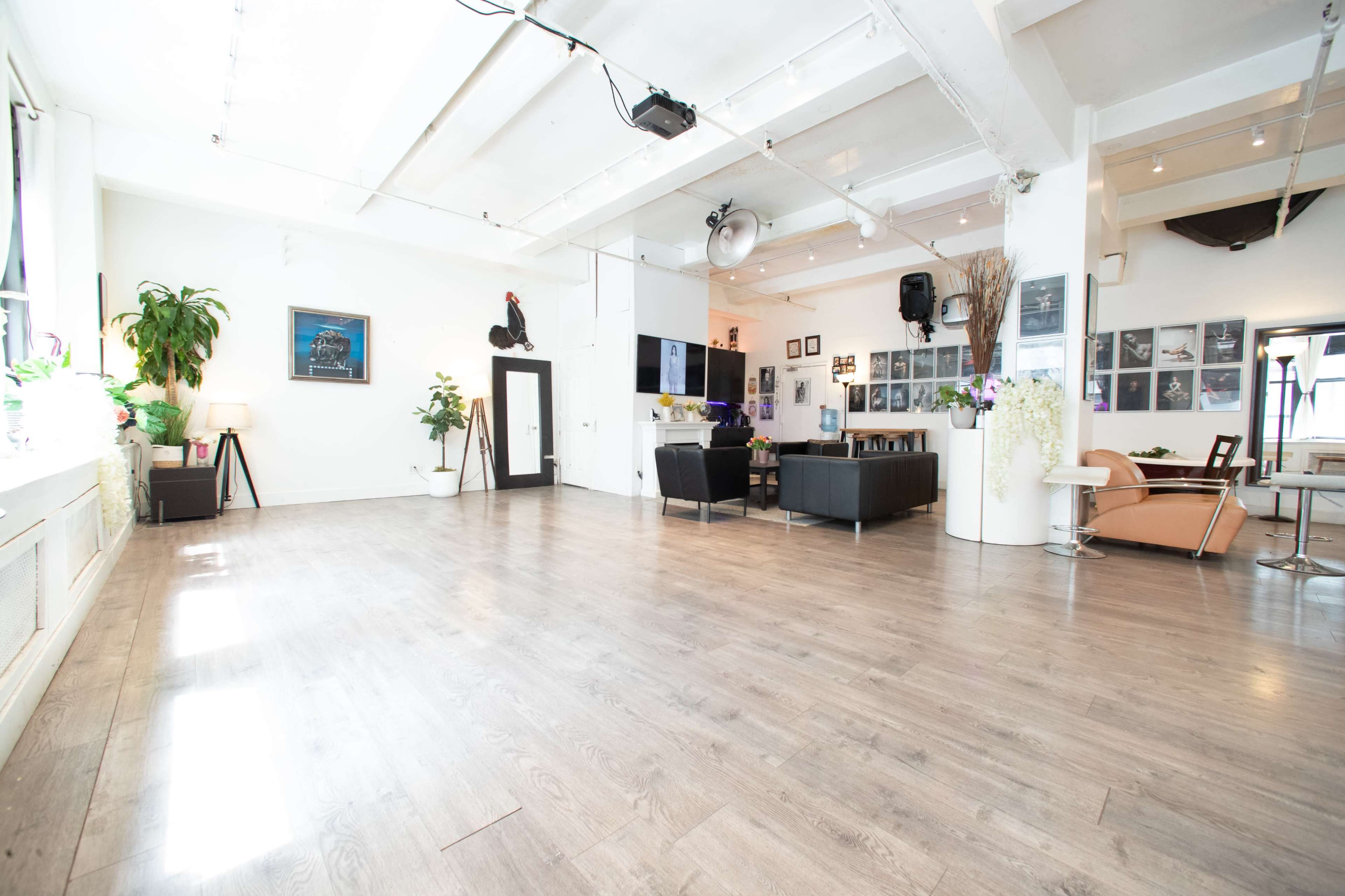 The image depicts a spacious, well-lit room with wooden flooring, featuring a seating area, plants, and walls adorned with photographs.