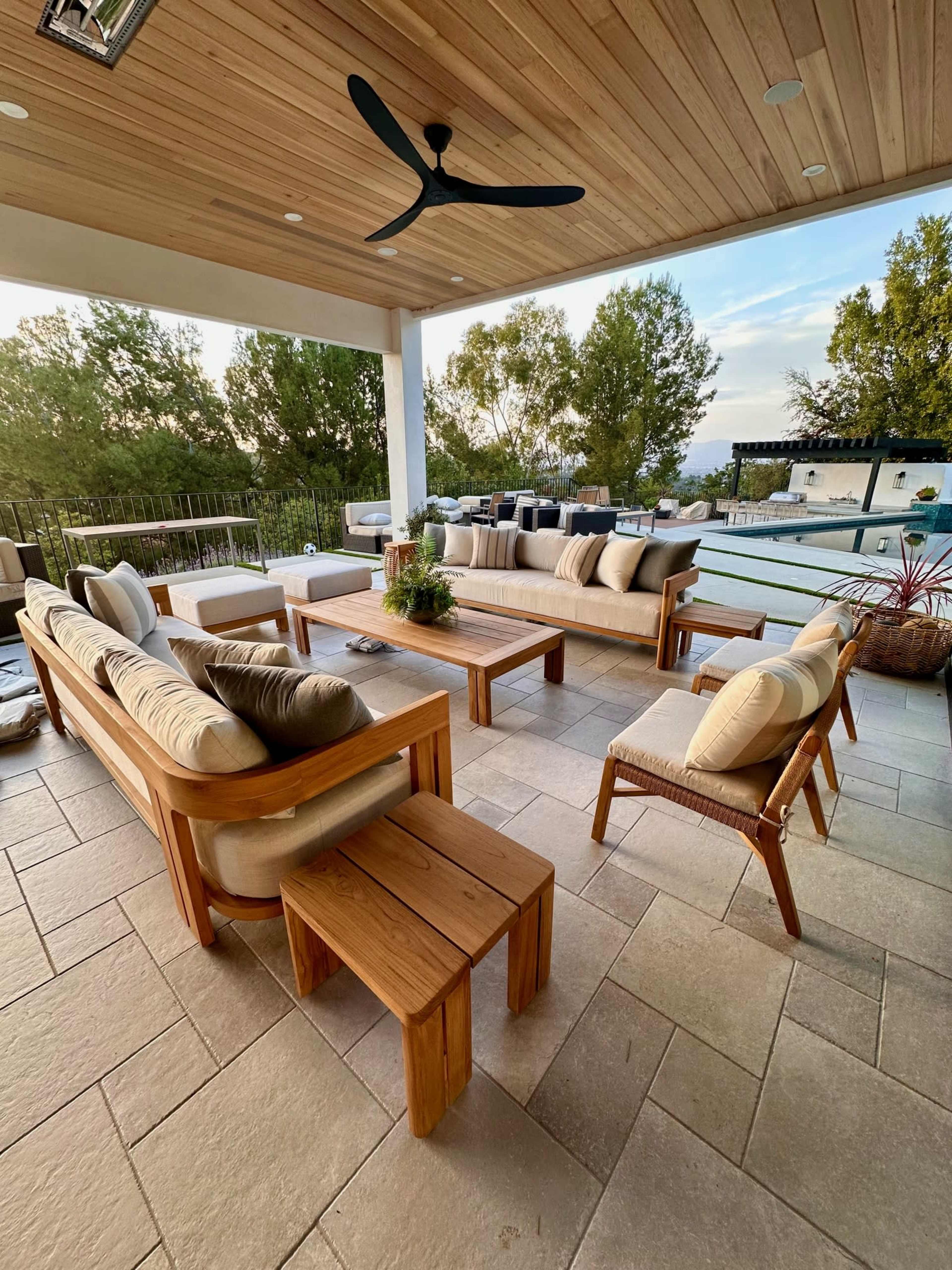 A spacious outdoor lounge area features wooden furniture, including a large sectional sofa and coffee table, set beside a swimming pool and surrounded by greenery.