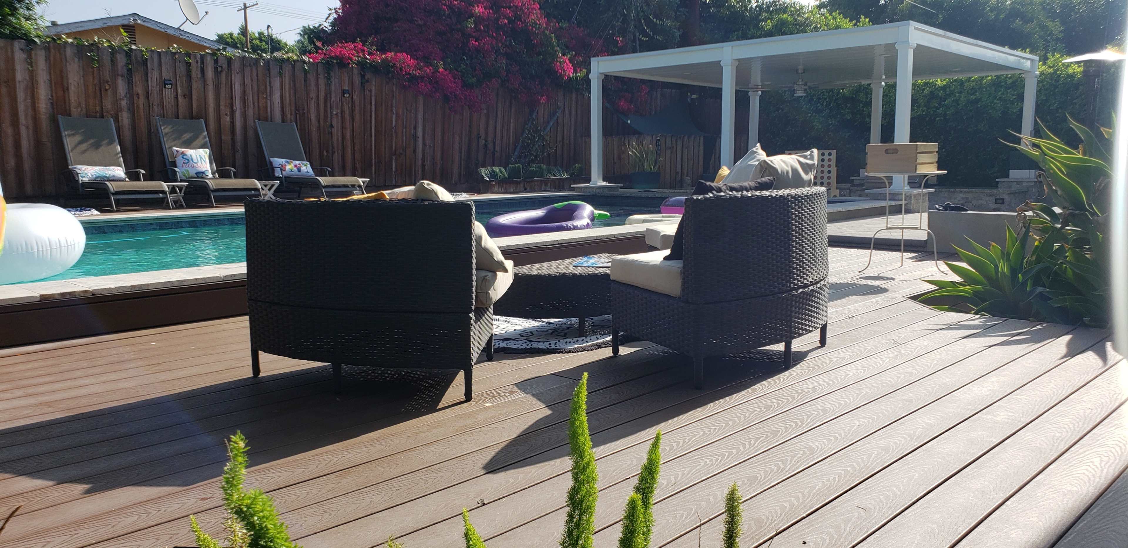 A backyard patio features two wicker chairs, a small table, and a swimming pool surrounded by greenery and a shaded lounging area.