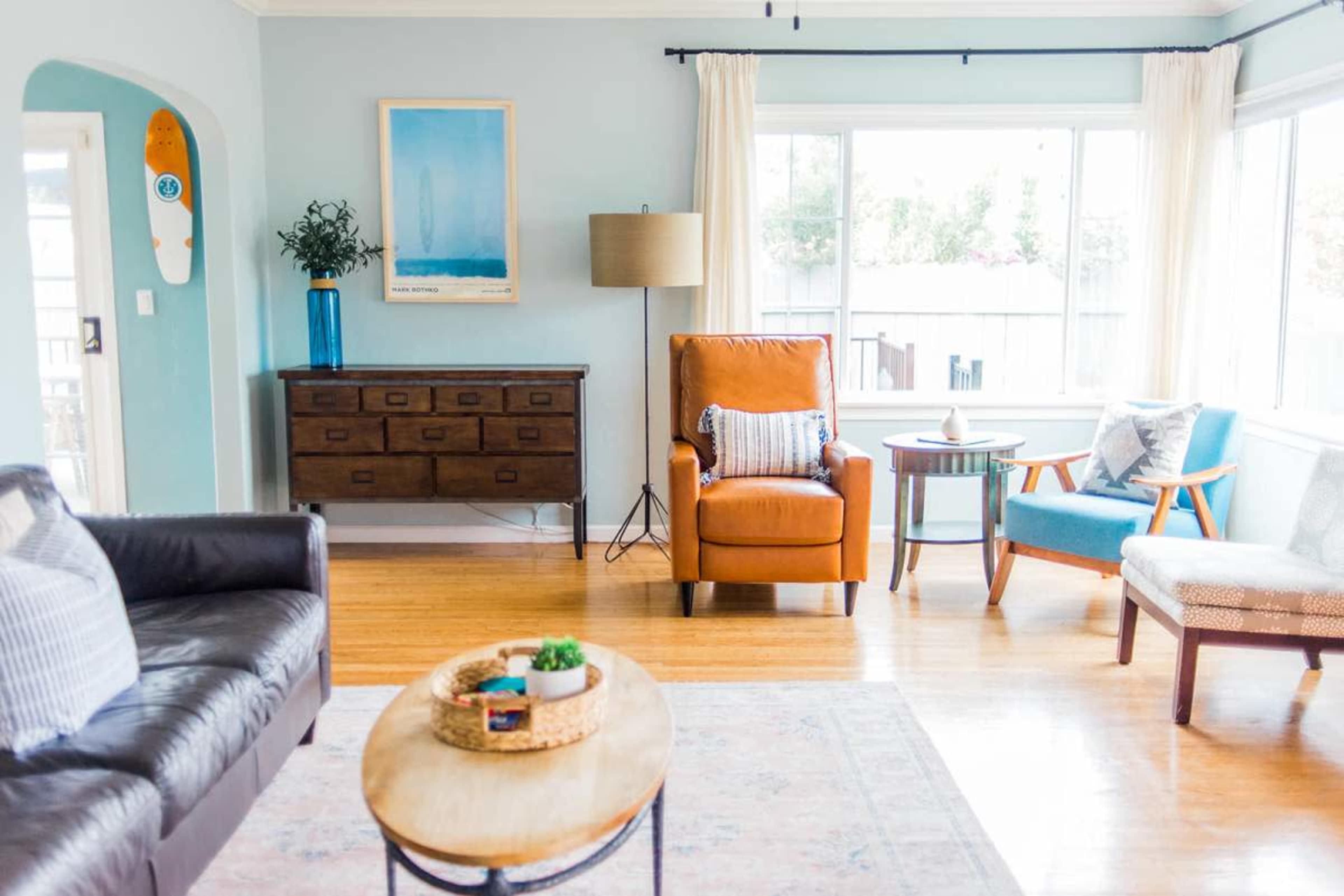 The image shows a cozy living room featuring a leather sofa, an orange armchair, a wooden side table, and large windows that bring in natural light.
