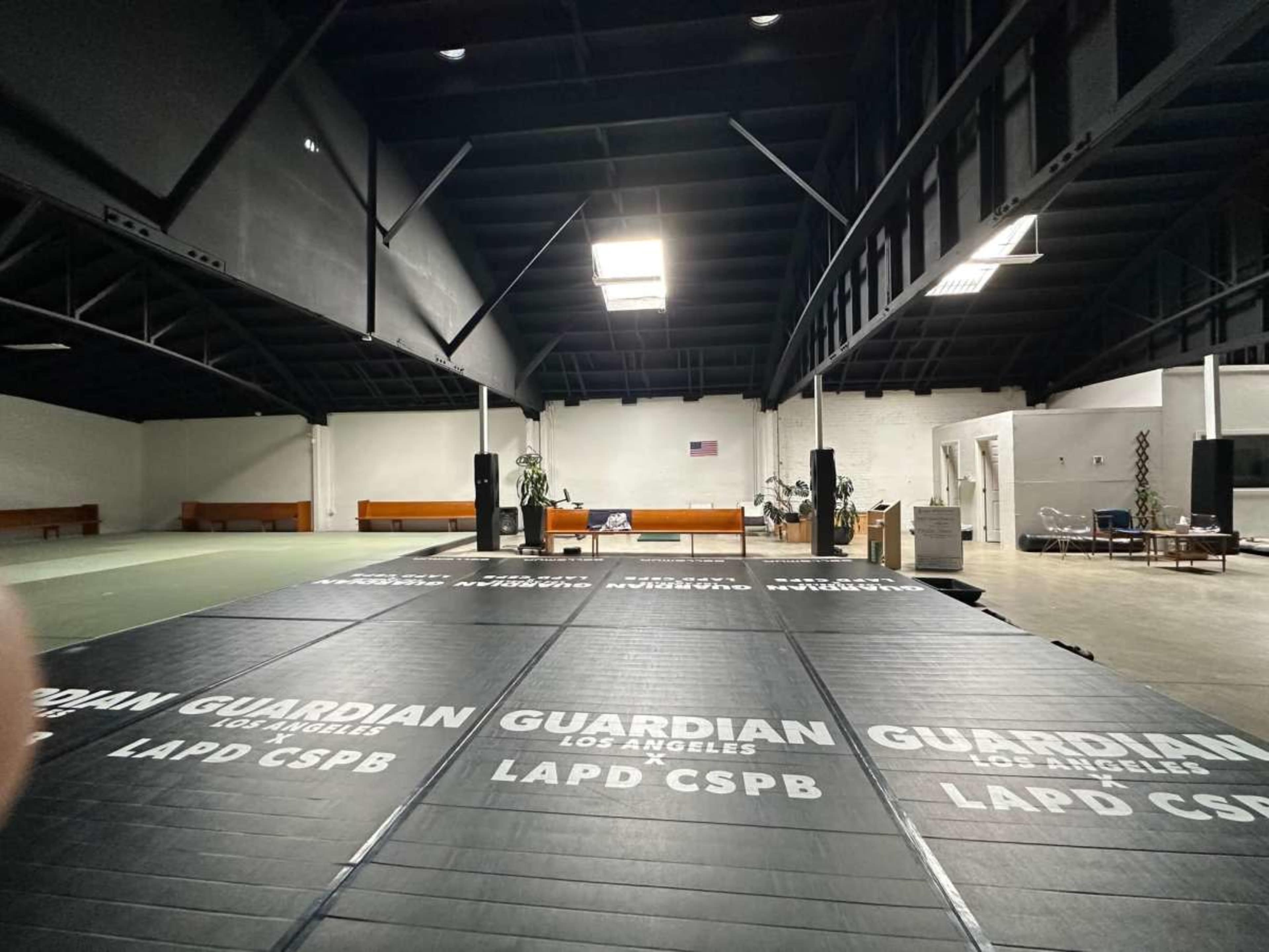 The image shows an empty training space featuring black mats with "GUARDIAN LOS ANGELES LAPD CSPB" printed on them, under a high ceiling with exposed beams.