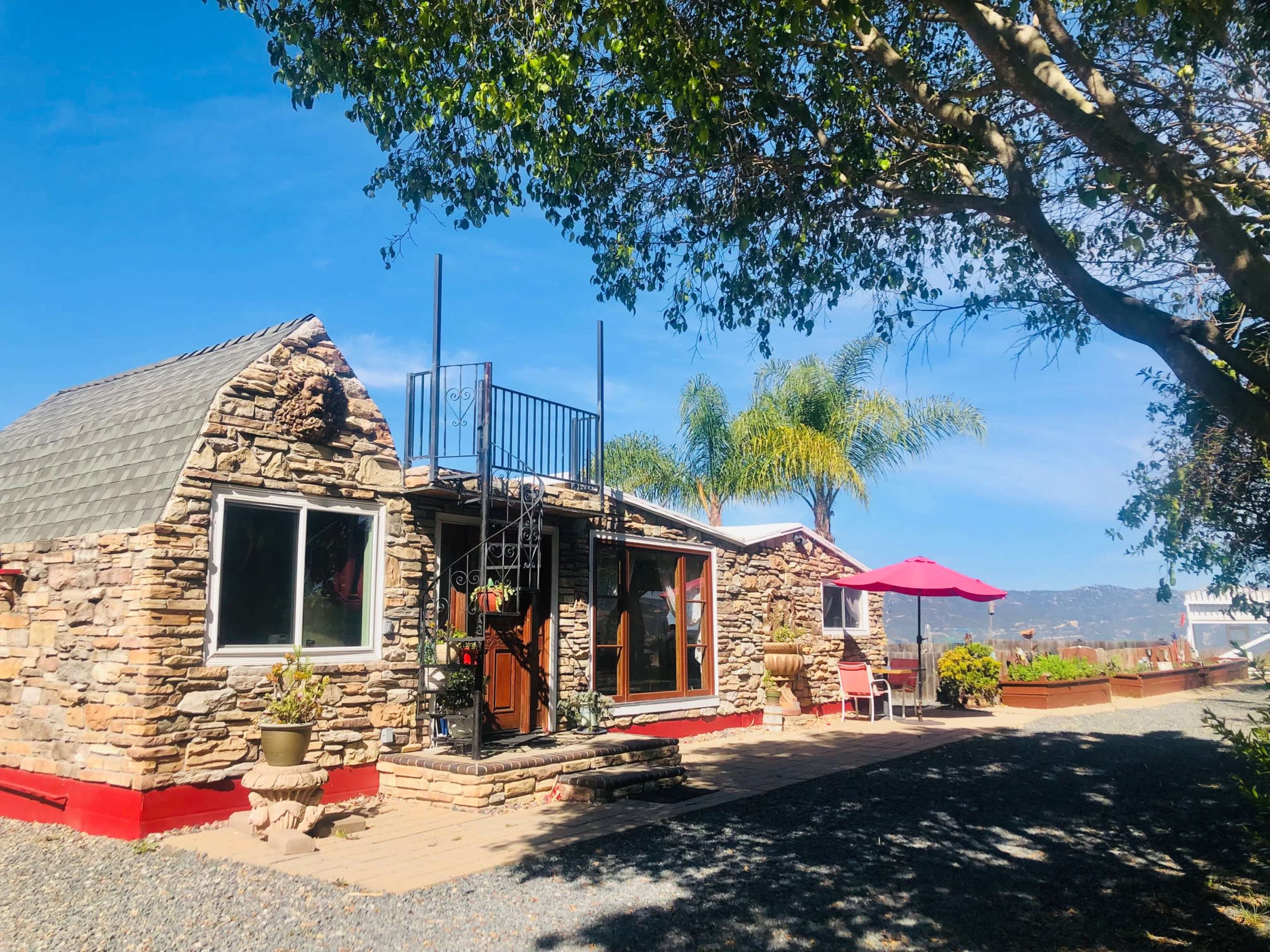 A stone cottage with a staircase leading to a rooftop deck is surrounded by palm trees and outdoor seating areas.