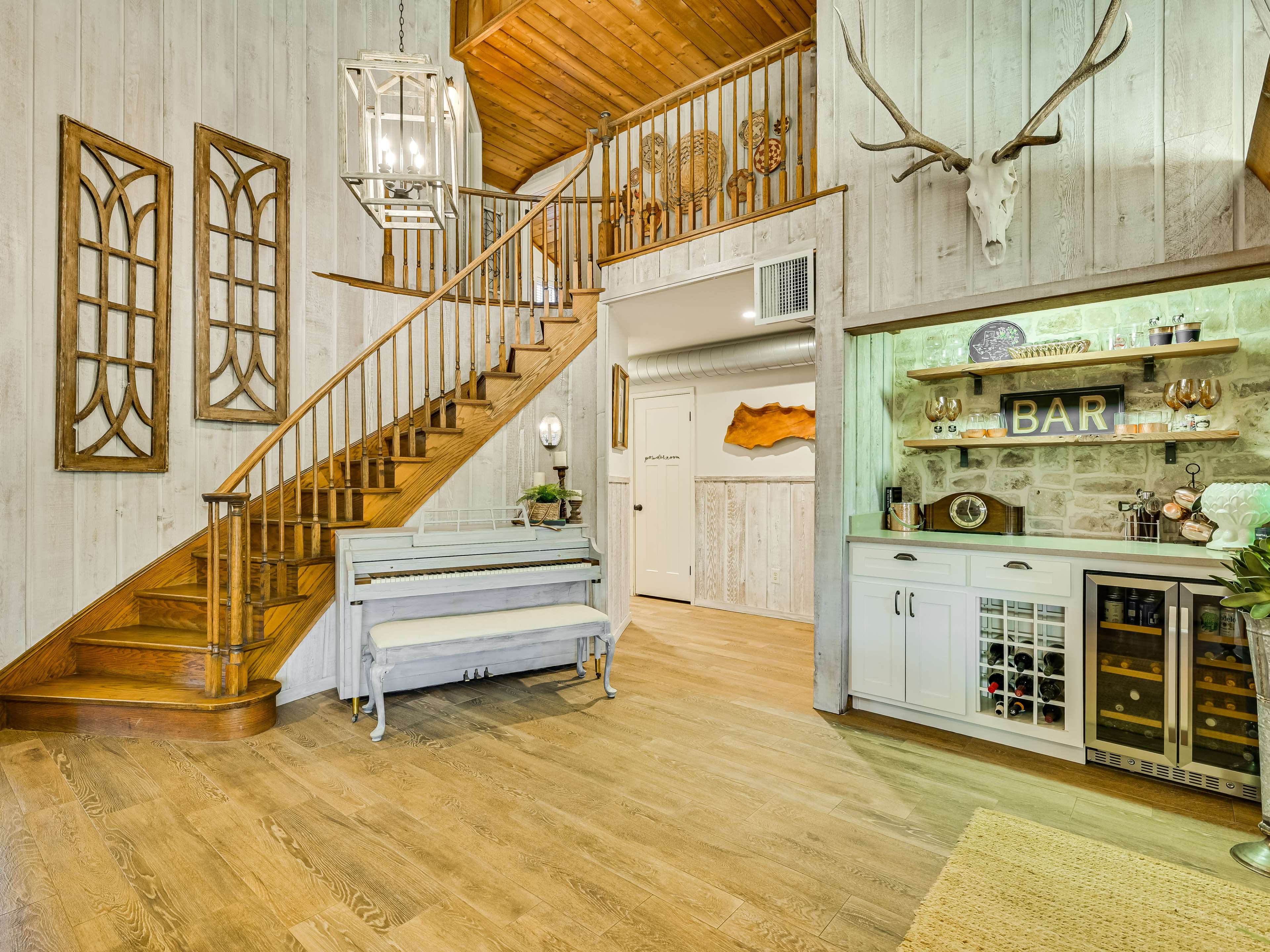 The image shows a well-decorated entryway featuring a wooden staircase, a vintage-style piano, and a bar area against a wooden wall.