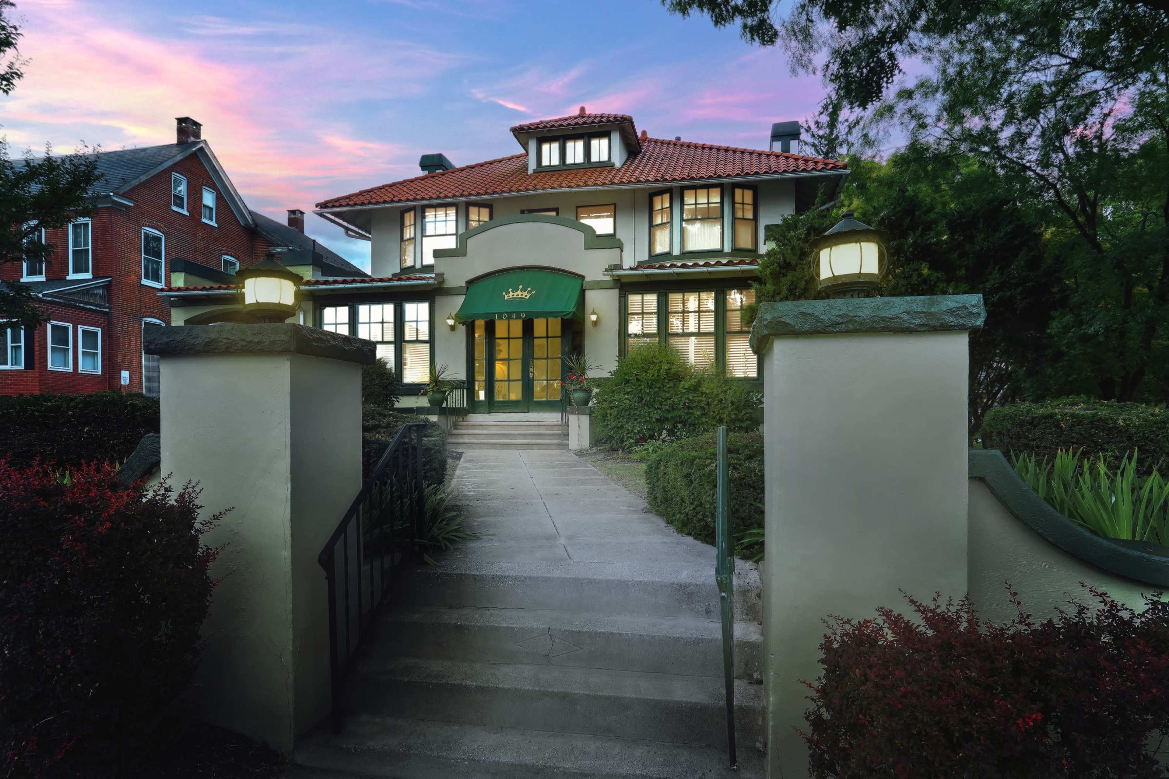 A two-story house with a green entrance and landscaped front steps is framed by greenery under a colorful sunset sky.