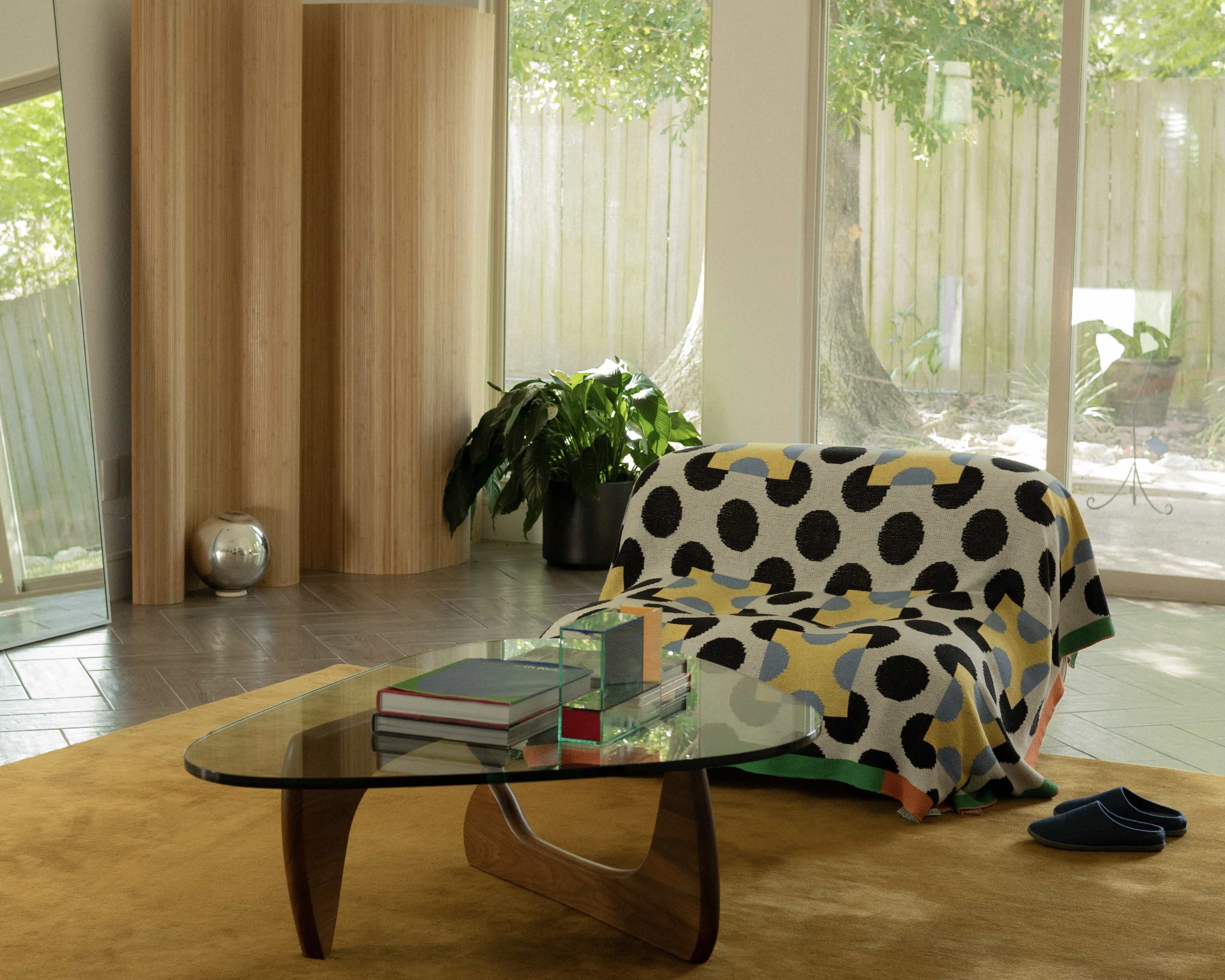 A cozy living room features a glass coffee table, a polka-dot patterned throw draped over a sofa, and large windows overlooking greenery.