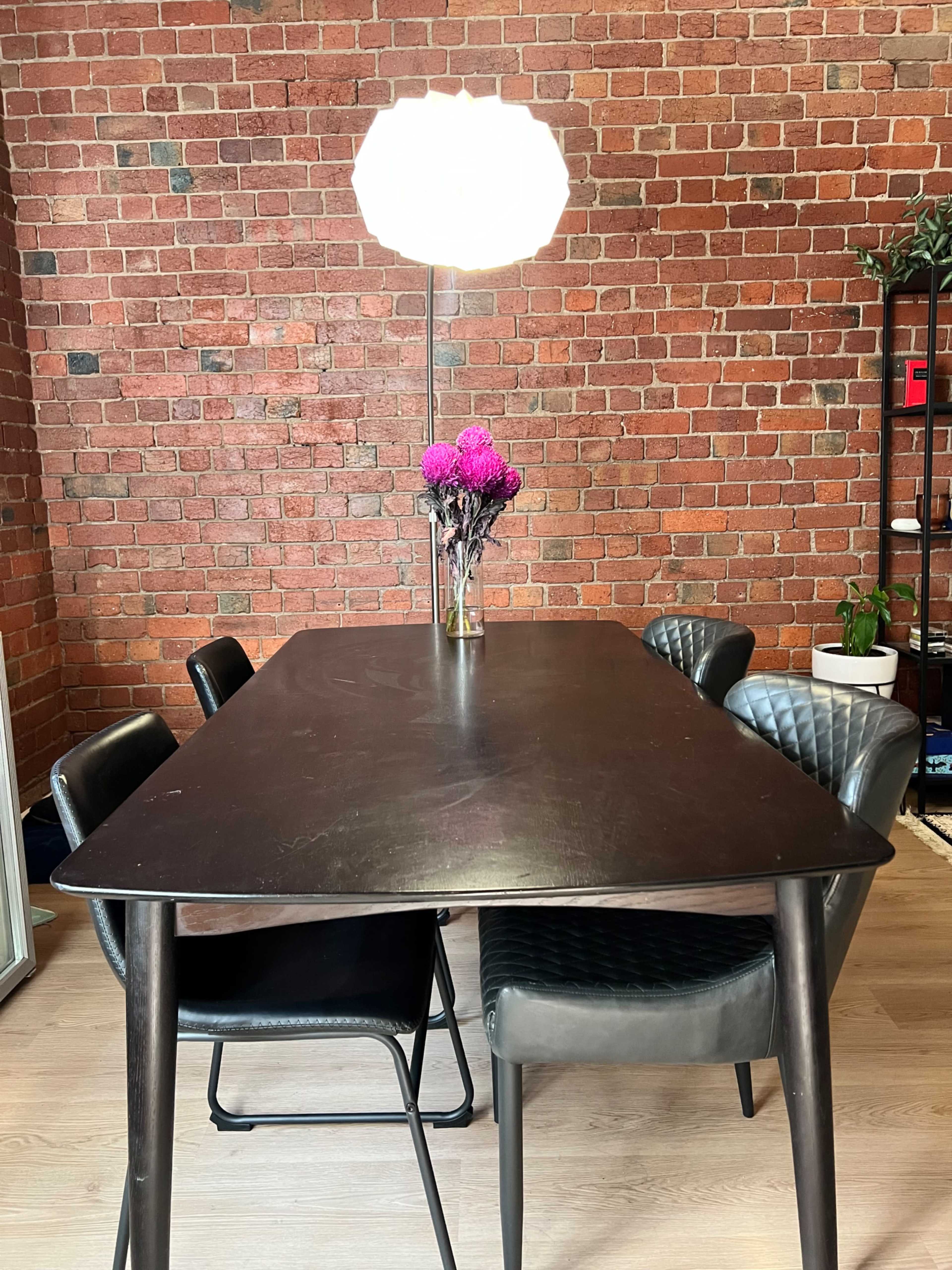 A black dining table with four chairs stands in a room with a brick wall, illuminated by a modern light fixture overhead and adorned with a vase of pink flowers.