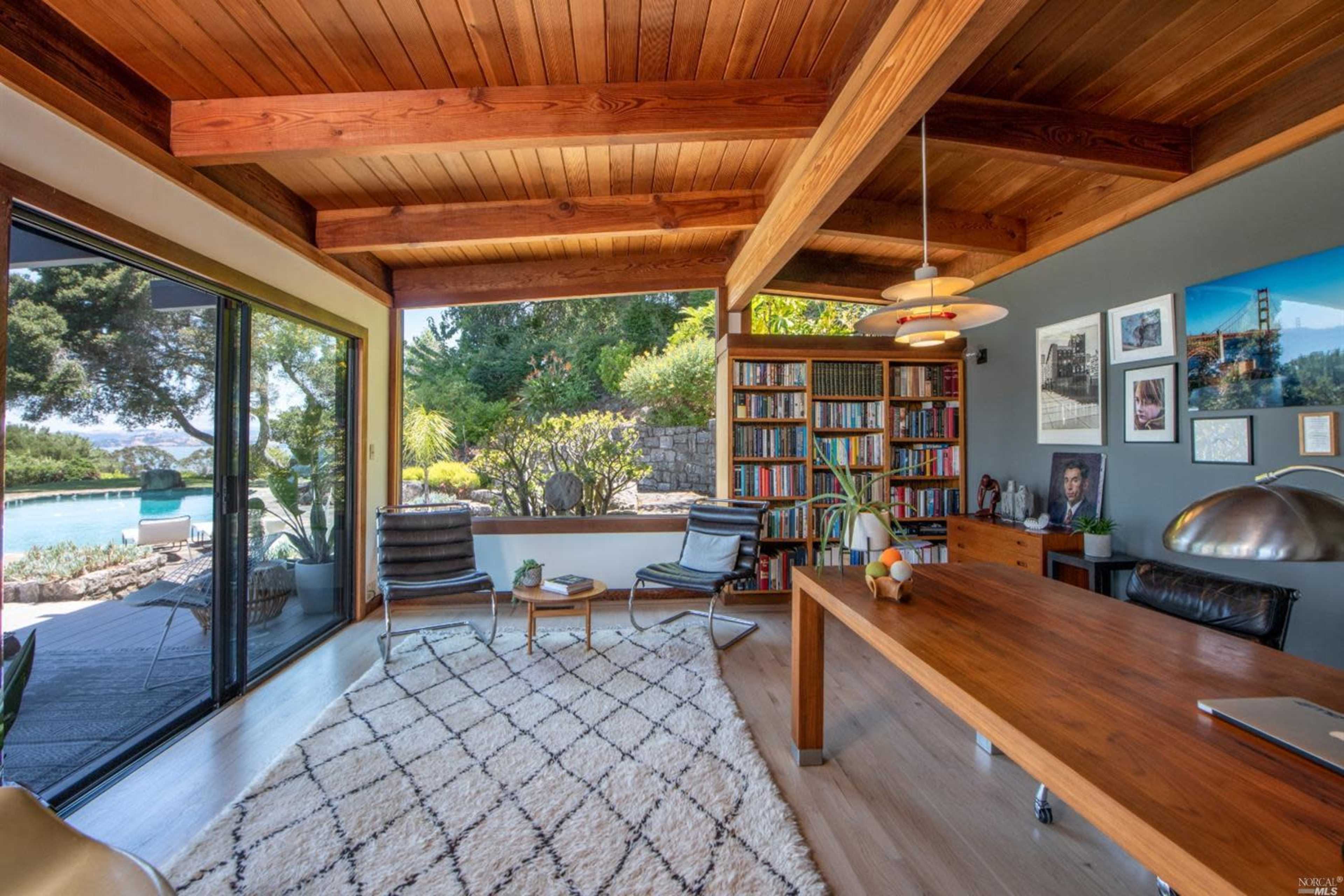 A modern home office with wooden beams, a large window overlooking a pool area, and a bookshelf filled with books.