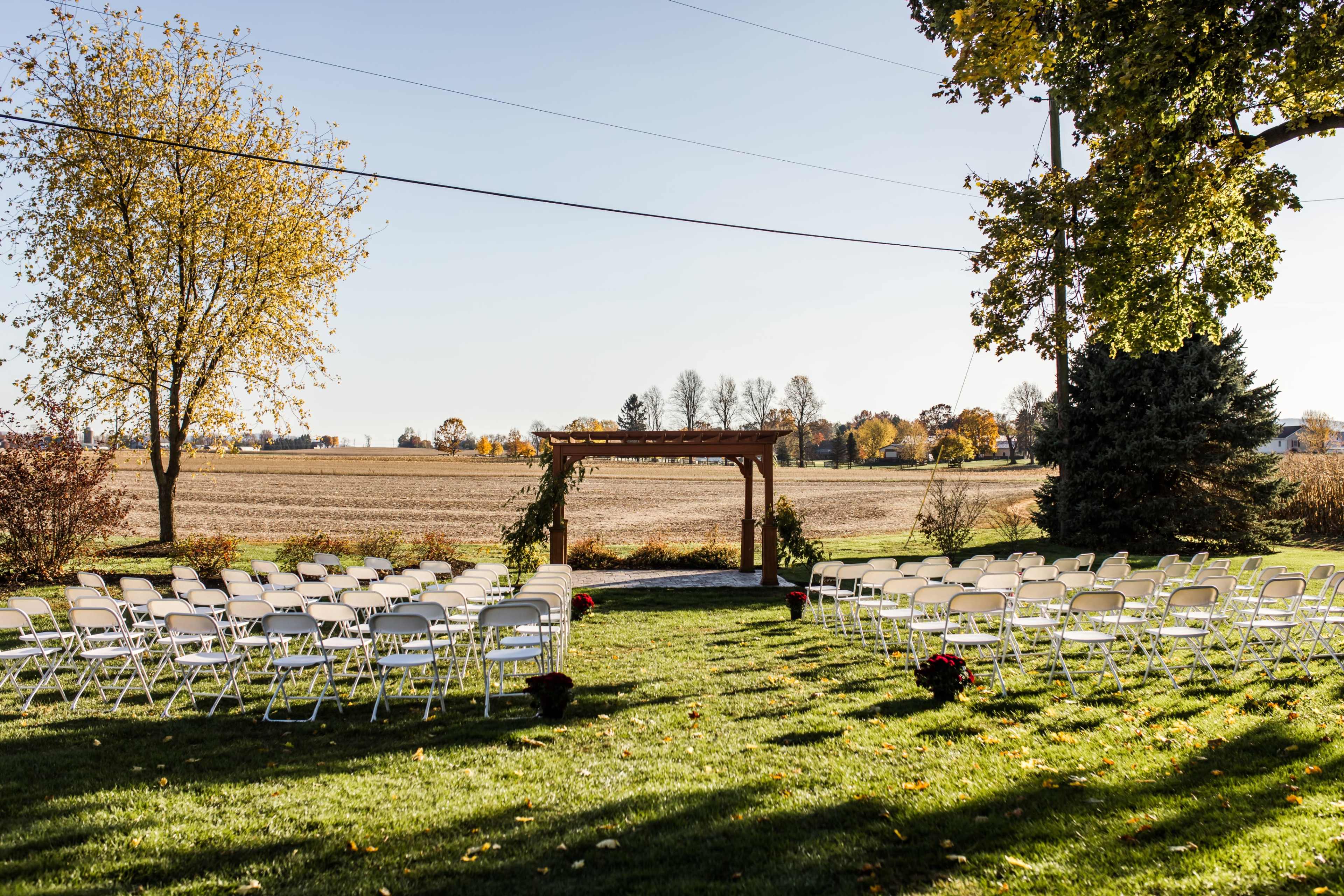 A wedding ceremony setup with rows of white chairs arranged on a grassy area in front of a wooden arbor, overlooking a field and trees in the background.