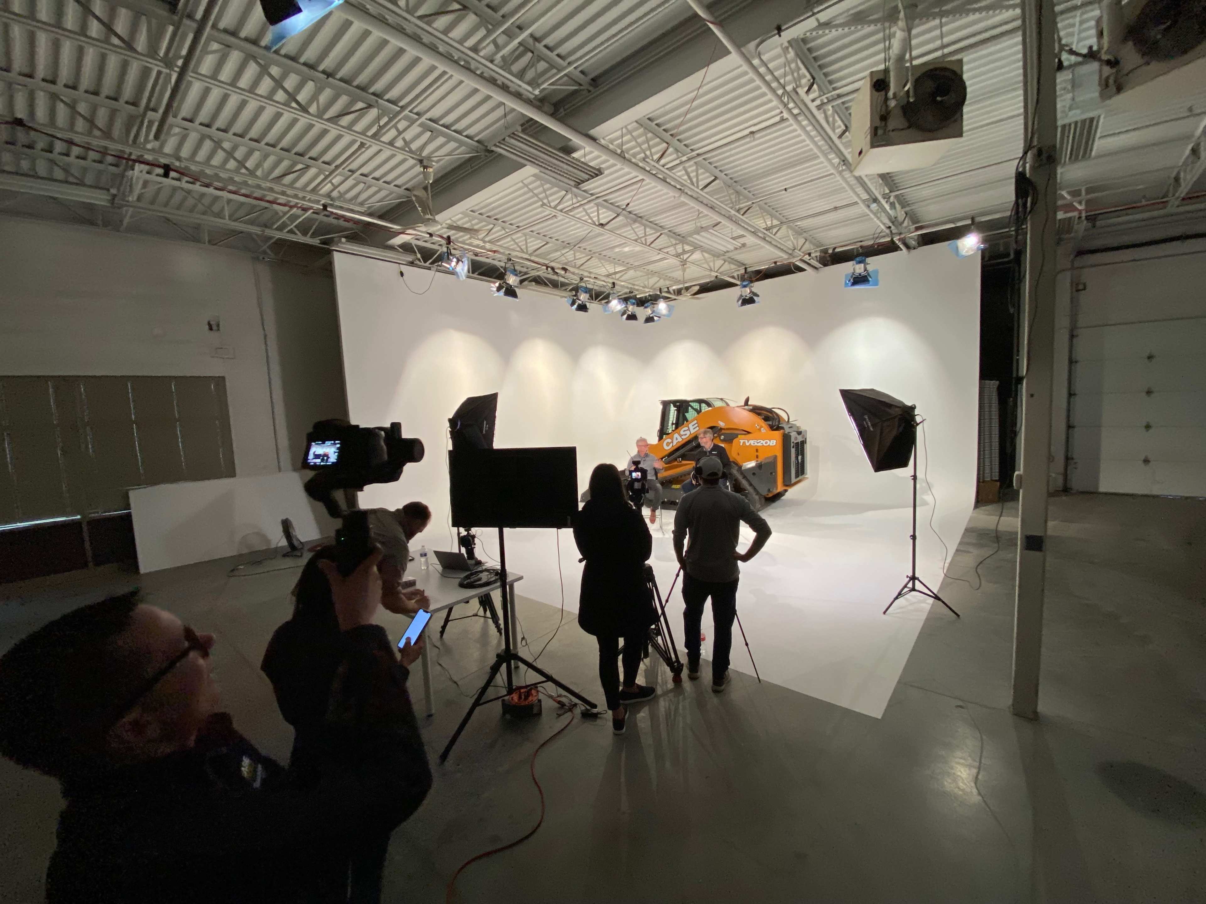 A group of people is setting up equipment in a studio space for a photo shoot featuring a large orange piece of machinery.
