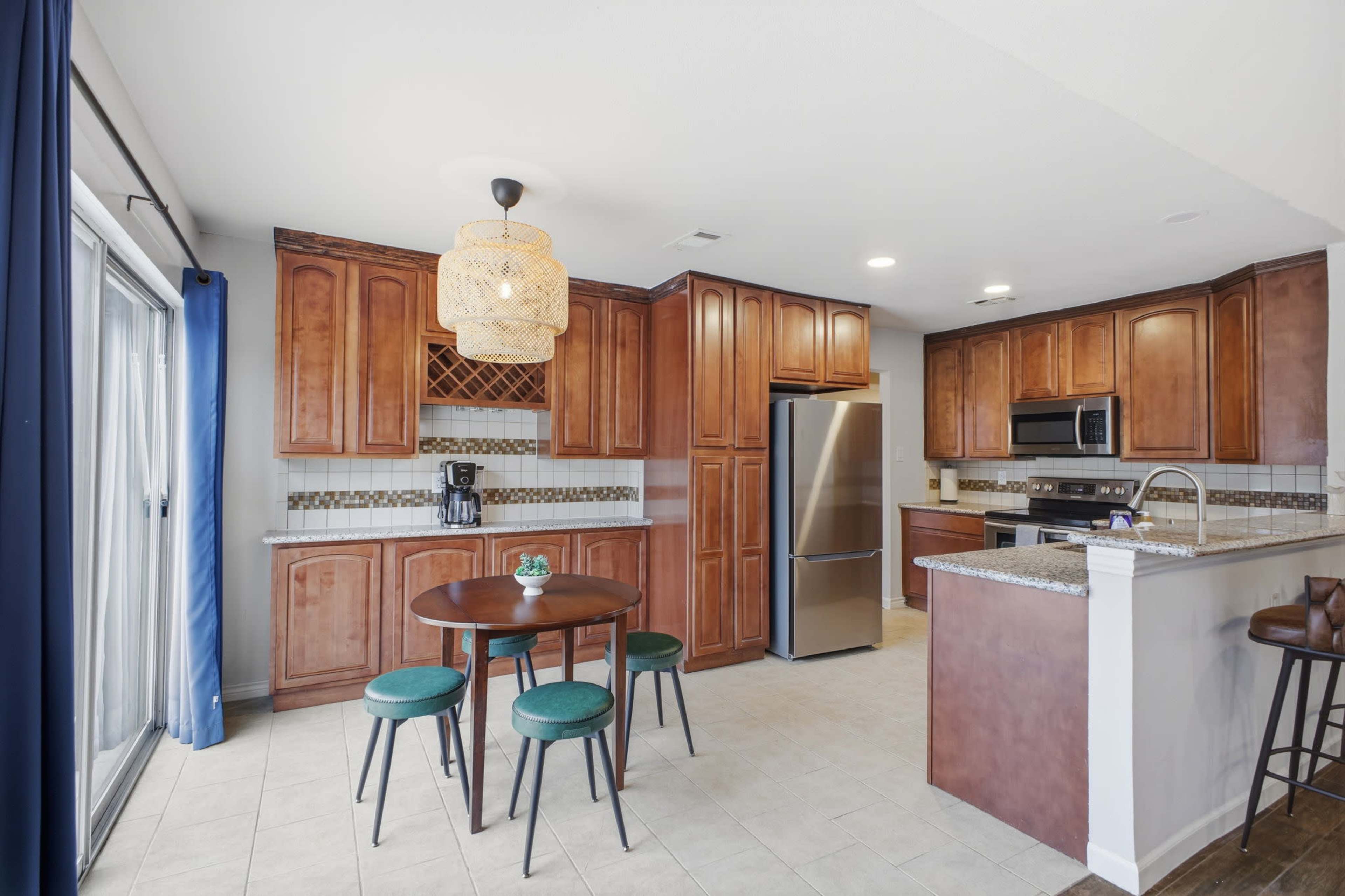 A spacious kitchen features wooden cabinets, a silver refrigerator, and a round dining table with four green stools.