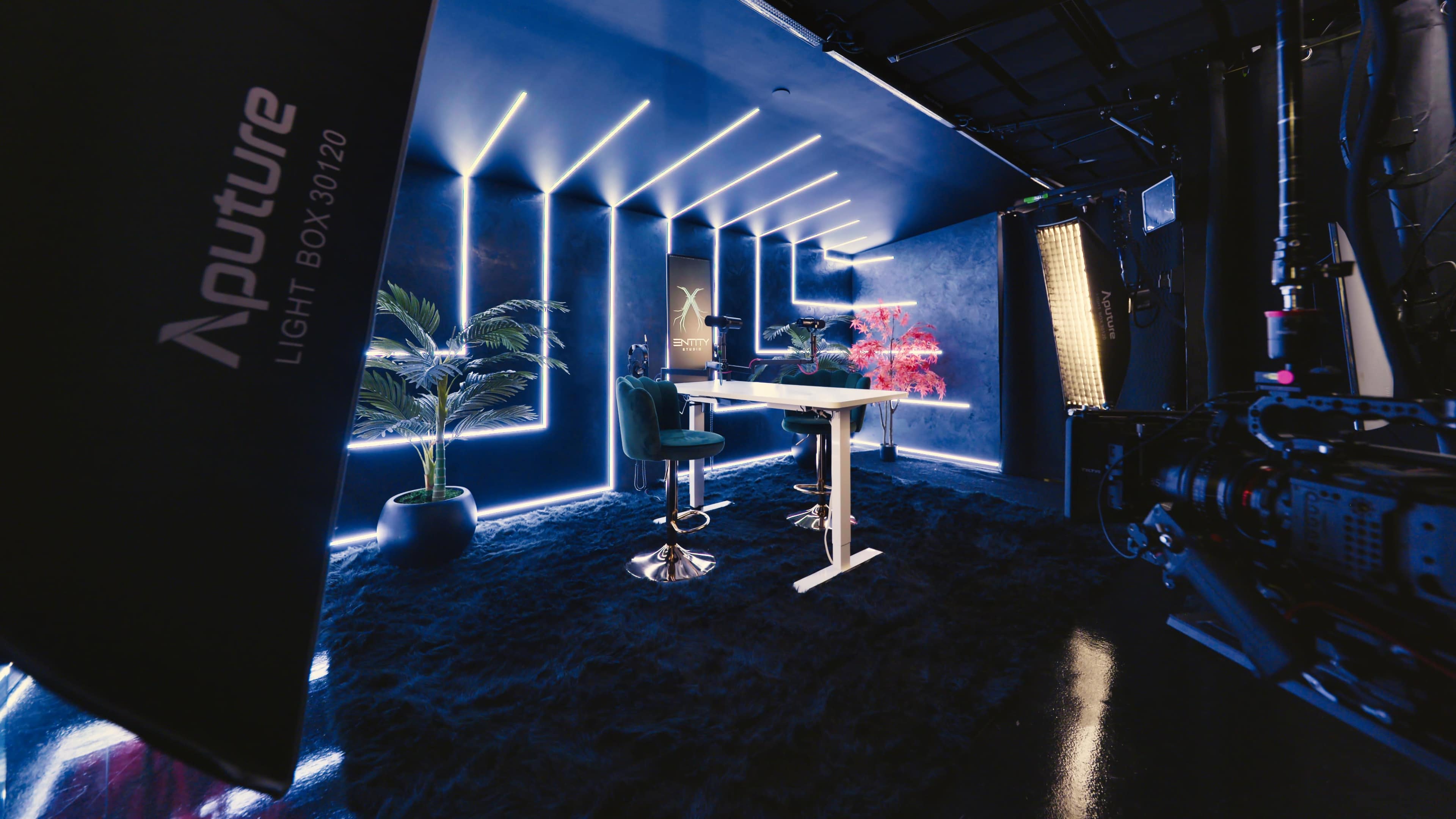 The image shows a modern studio space with blue walls, neon lighting, a white table with two green chairs, and a large rug.