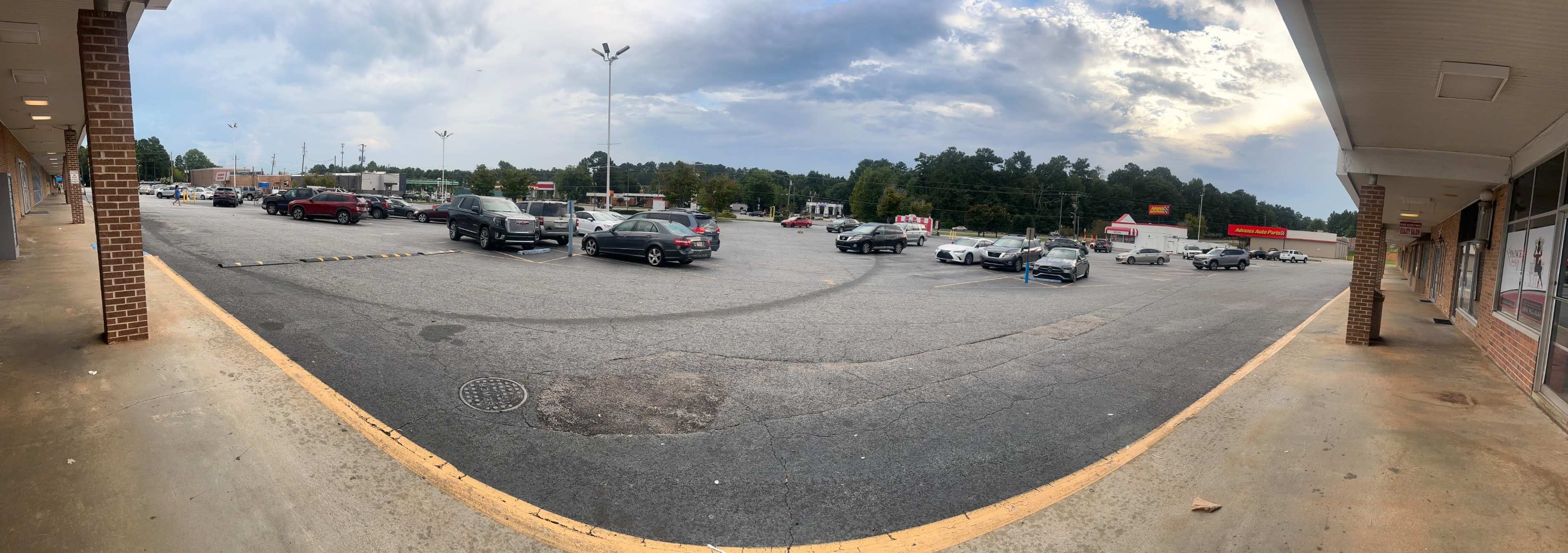 The image shows a wide view of a partially filled parking lot in front of a strip mall, with various cars parked and clouds in the sky.